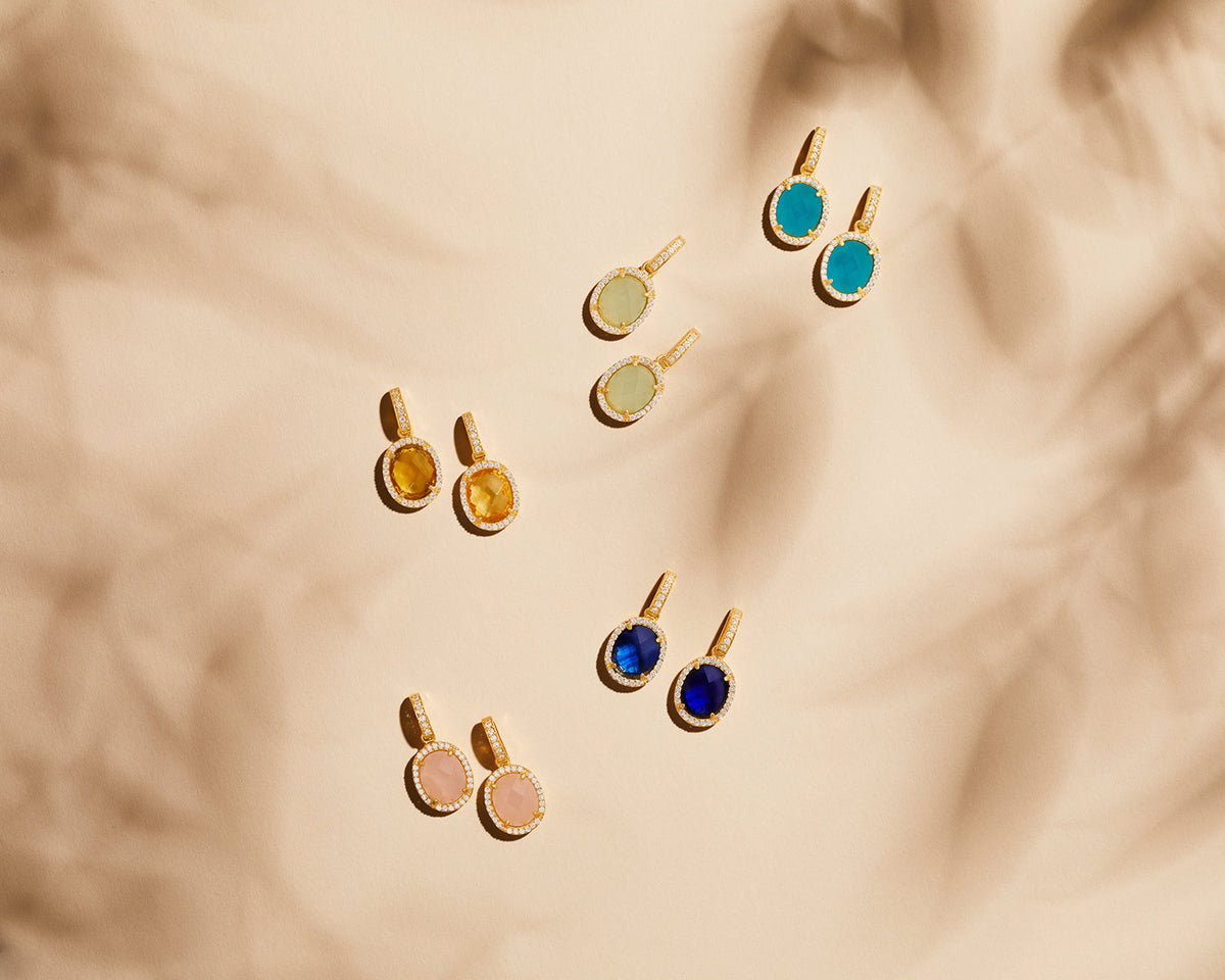 Assortment of colorful oval gemstone pendants with gold diamond halos on a beige background