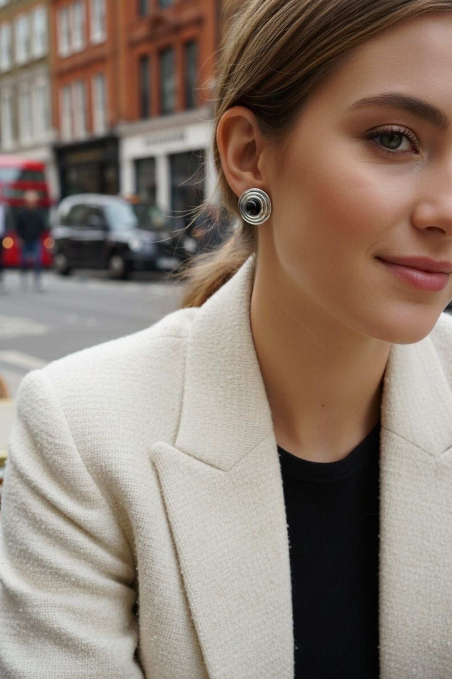 Woman in a beige blazer with a blurred city street background wearing a silver earring with a black centre