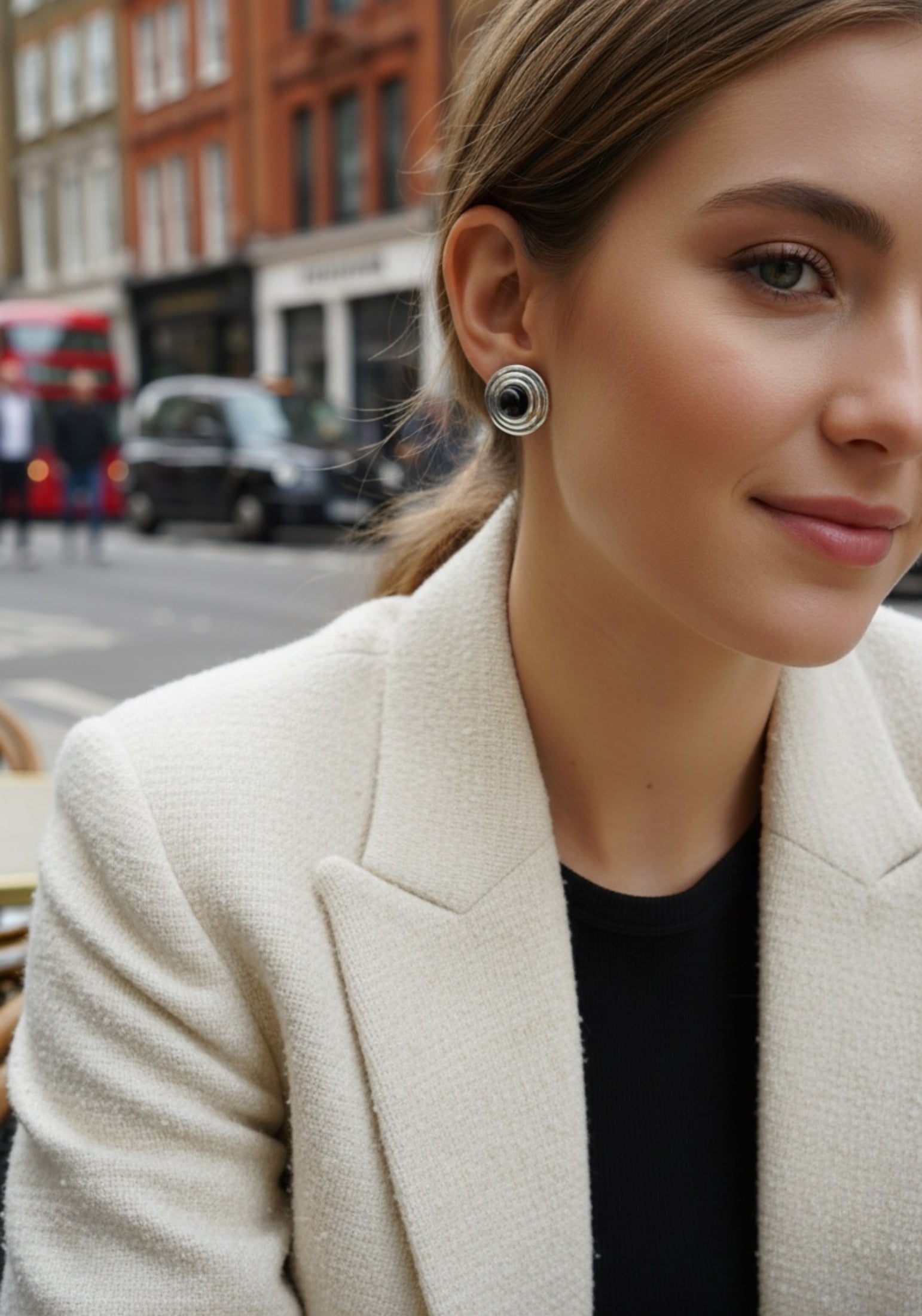 Woman in a beige blazer with a blurred city street background wearing a silver earring with a black centre
