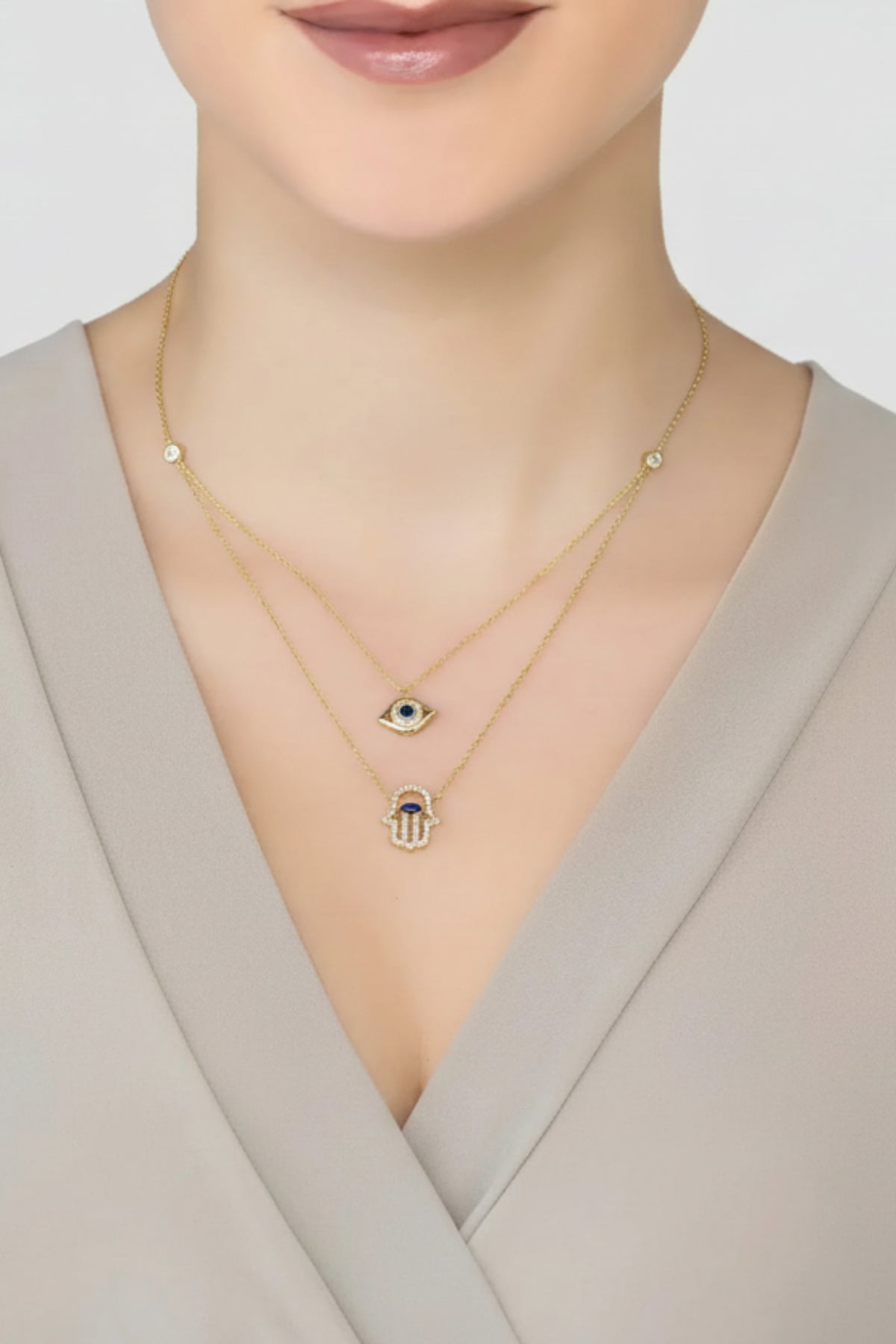 A close-up of a model wearing a double-layered gold necklace set. The top layer features a small gold hamsa hand pendant, and the bottom layer features an evil eye charm with a green and gold center.