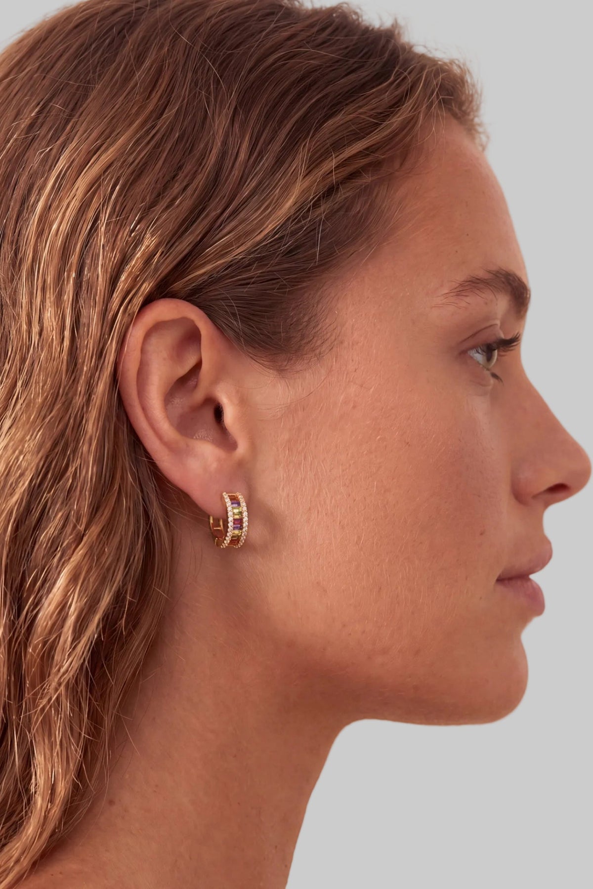 A close-up side profile of a woman wearing a small gold hoop earring adorned with multiple rows of colorful rhinestones.