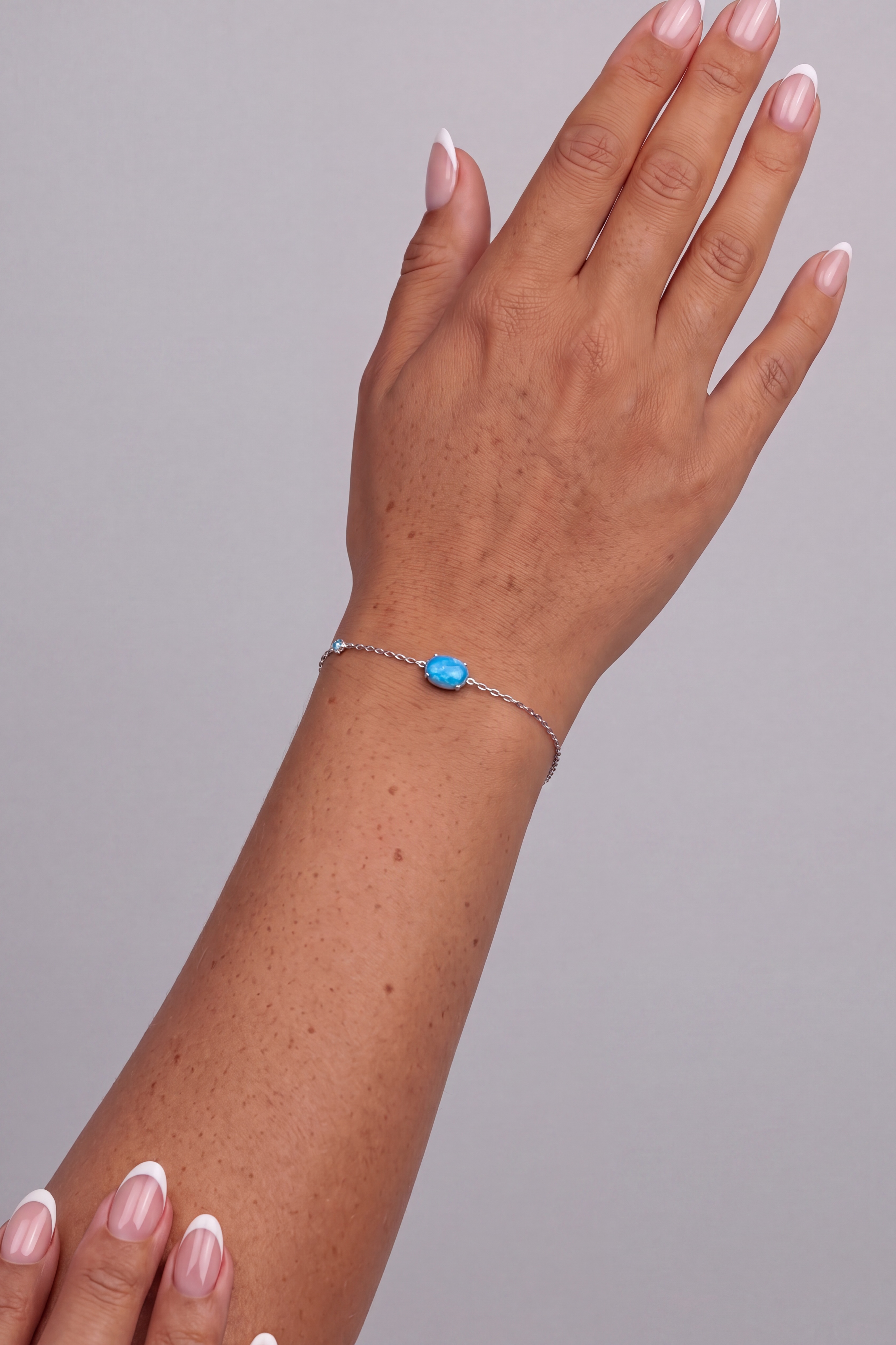 A delicate silver chain bracelet with a small oval blue stone, shown worn on a woman's wrist. The model's hand is posed against a solid neutral gray background.