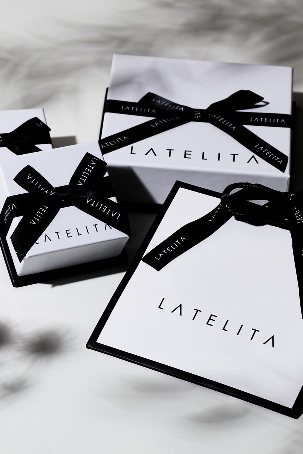 White LATELITA gift boxes and a matching shopping bag tied with black ribbon, arranged on a light marble surface with soft shadows.