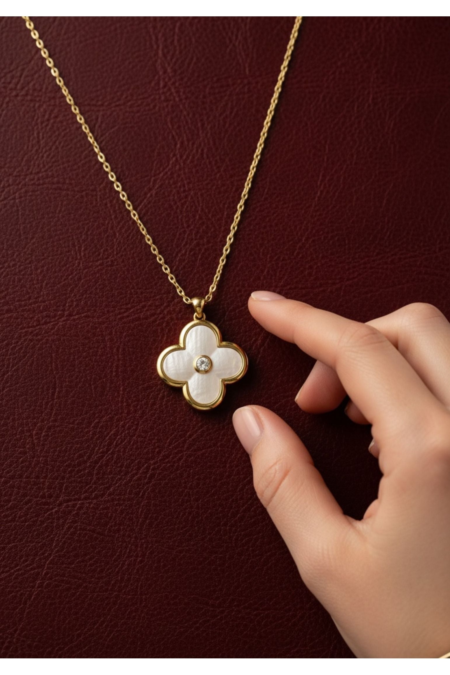 Gold necklace with a clover-shaped pendant held by a hand against a dark red background