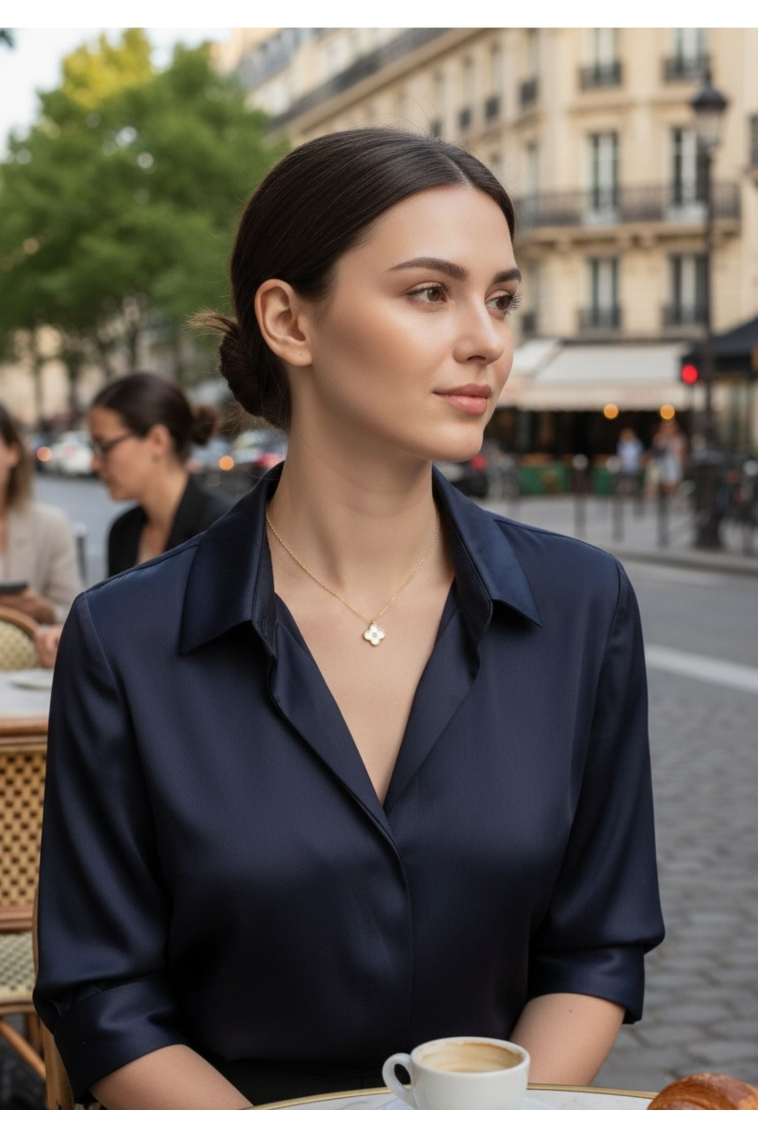 Woman in a navy blue shirt sitting at an outdoor cafe table with a city street background wearing a necklace with a clover pendant