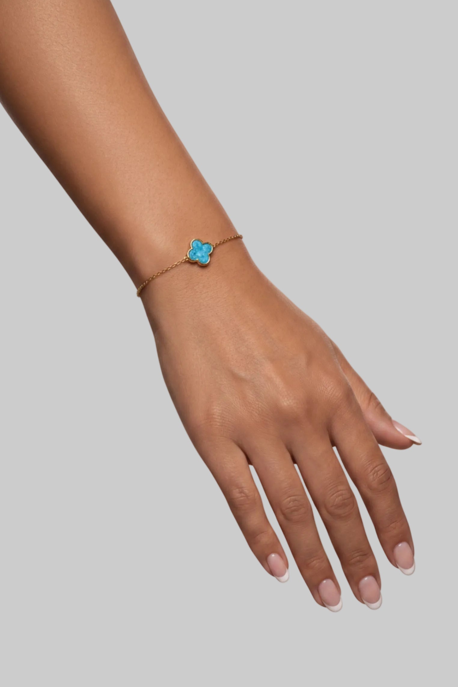 A delicate gold-tone bracelet with a small blue flower charm, worn on a model's wrist resting on a textured gray block.