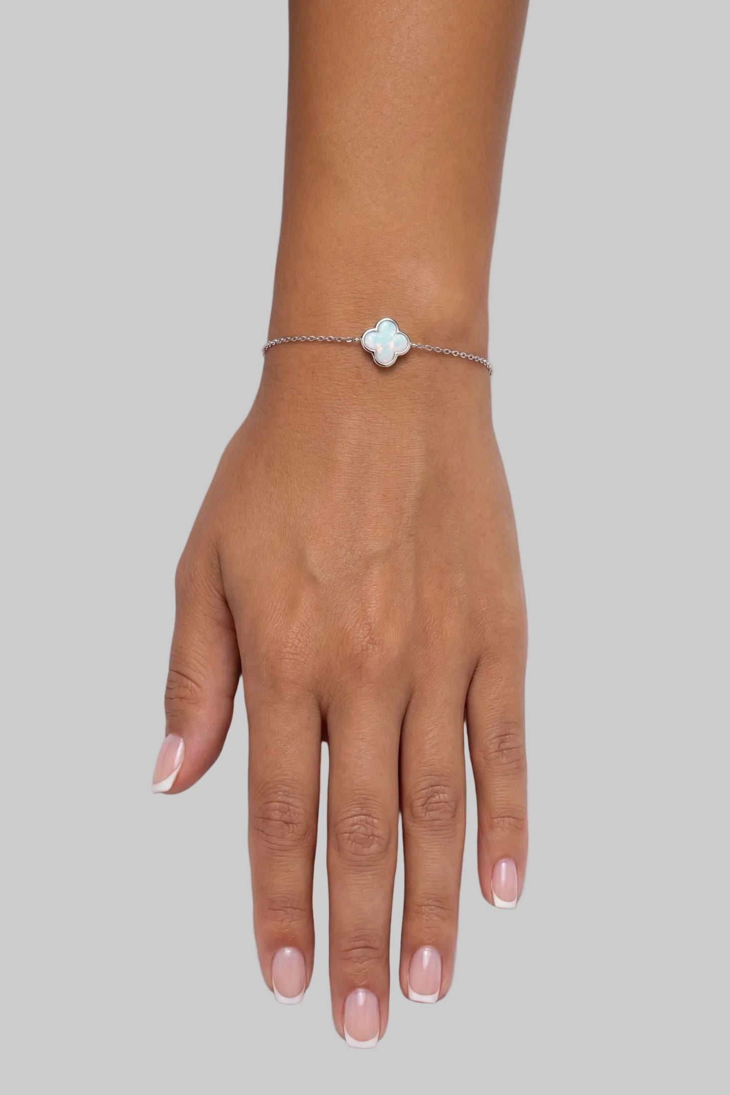A delicate silver chain bracelet with a small, light blue-green iridescent clover charm, shown worn on a person's wrist against a plain light gray background.