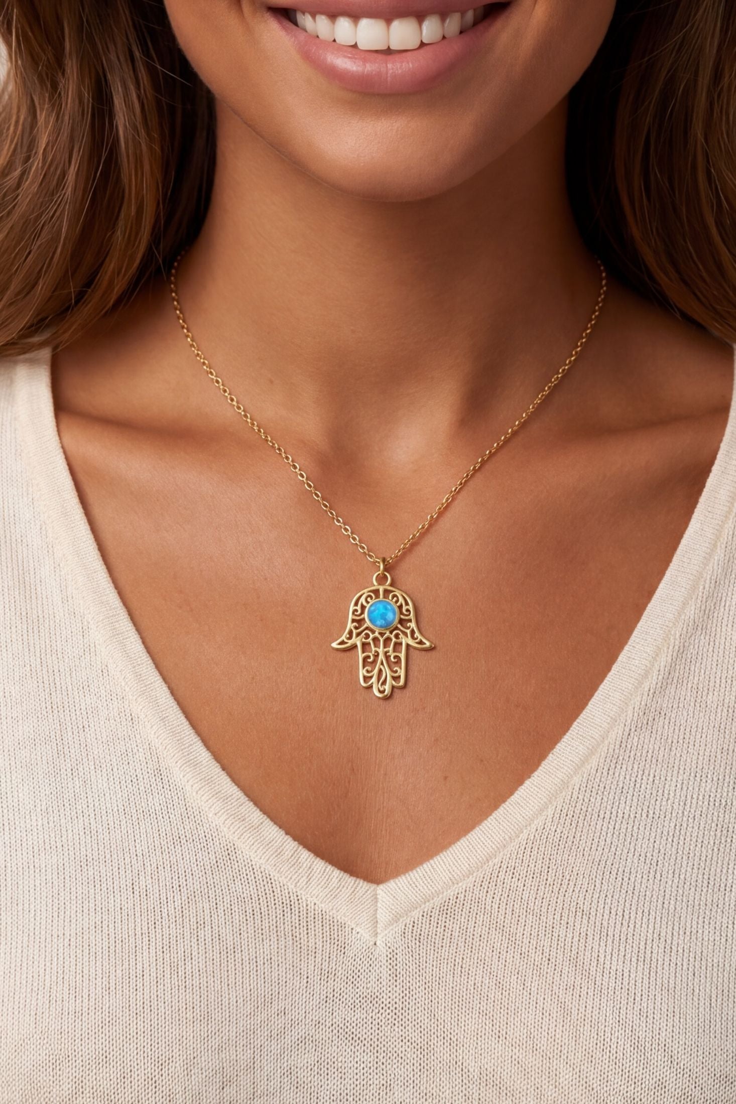 A gold Hamsa hand pendant necklace with a small blue stone in the center, worn by a model in a cream-colored ribbed V-neck top.