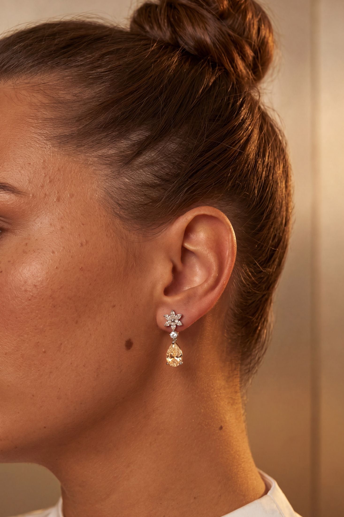 A close-up side profile of a woman with her hair in a bun, wearing a delicate gold dangle earring with a small floral stud and a teardrop-shaped clear crystal drop.
