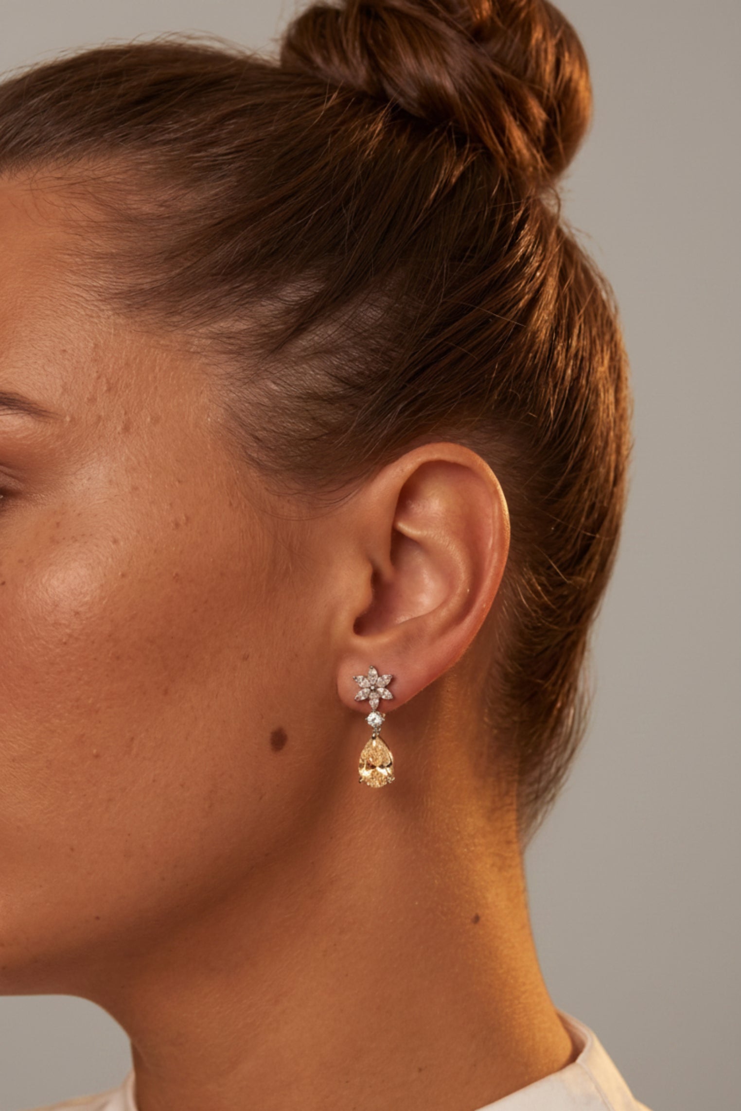 A close-up side profile of a woman with her hair in a bun, wearing a delicate gold dangle earring with a small floral stud and a teardrop-shaped clear crystal drop.