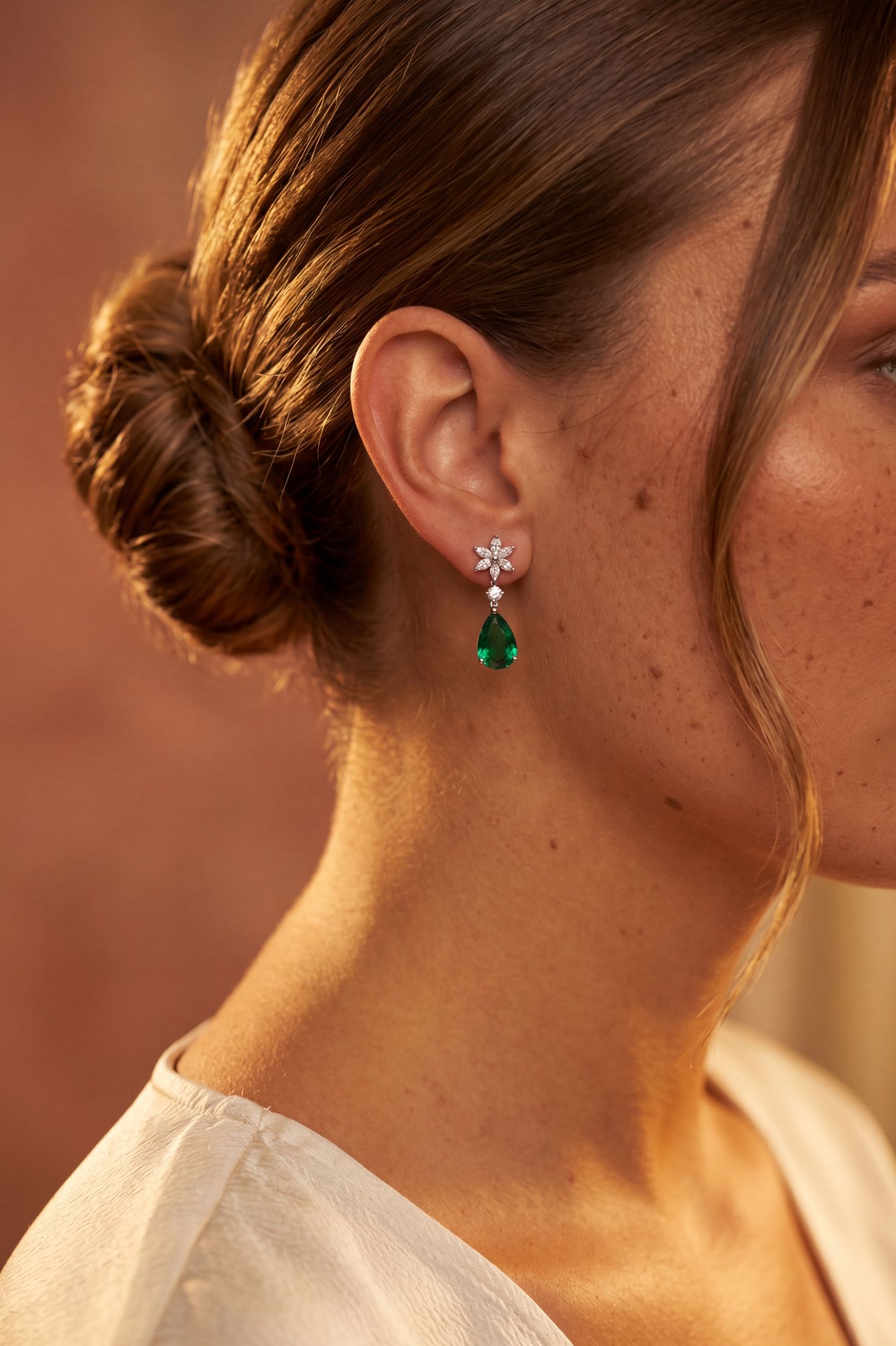 Close-up of a woman wearing a dangling earring with a silver-toned flower-shaped stud and a green teardrop gemstone, shown against warm brown skin tones and a soft brown background.