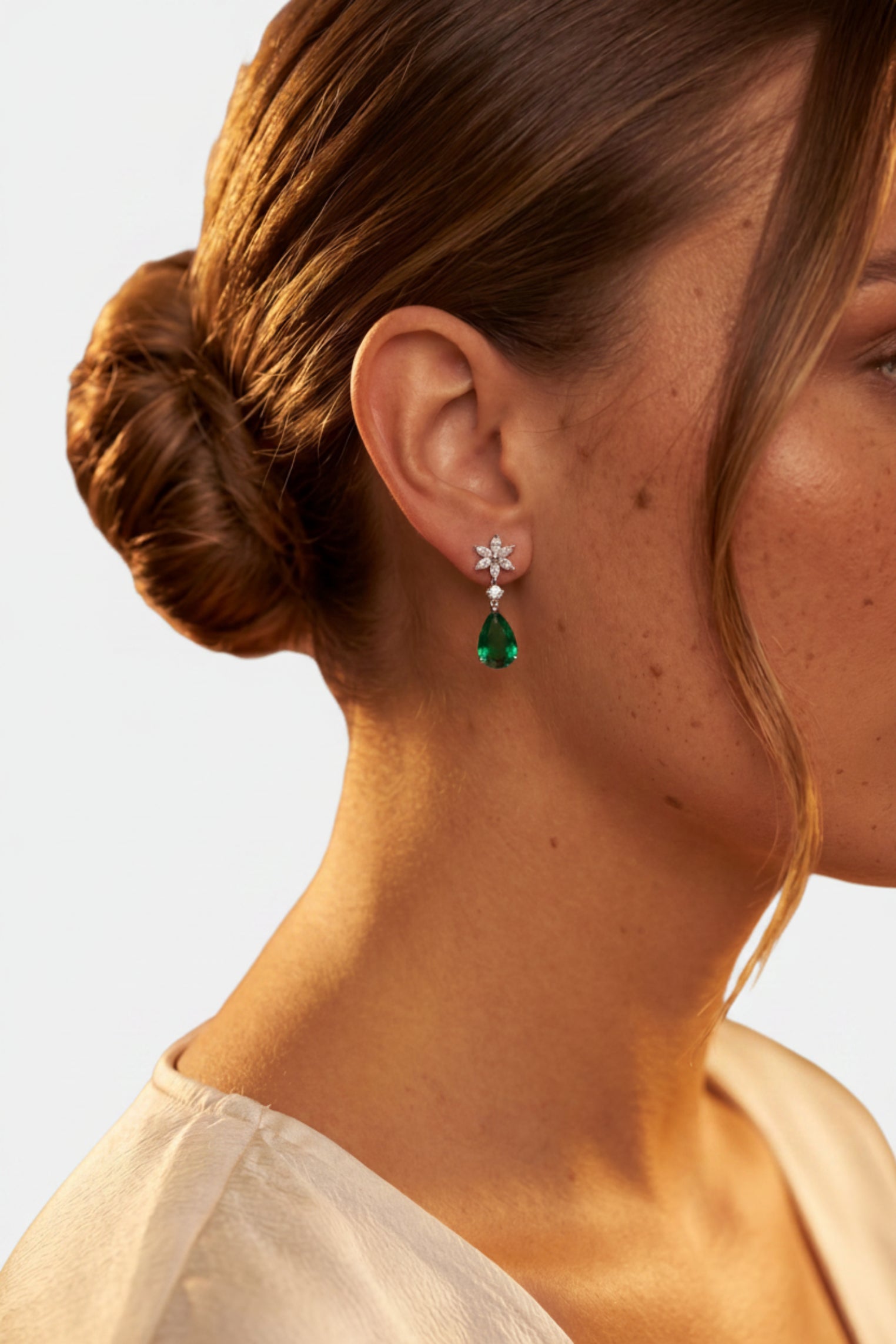 Close-up of a woman wearing a dangling earring with a silver-toned flower-shaped stud and a green teardrop gemstone, shown against warm brown skin tones and a soft brown background.