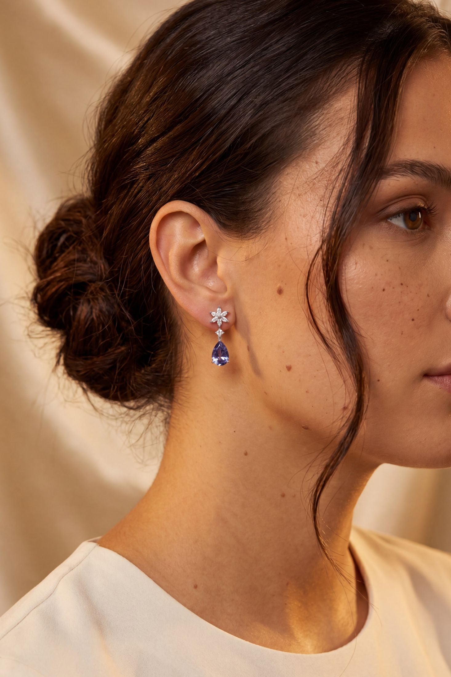 A close-up side profile of a woman wearing a delicate drop earring with a silver-toned floral stud and a deep blue teardrop pendant, set against a soft beige background.