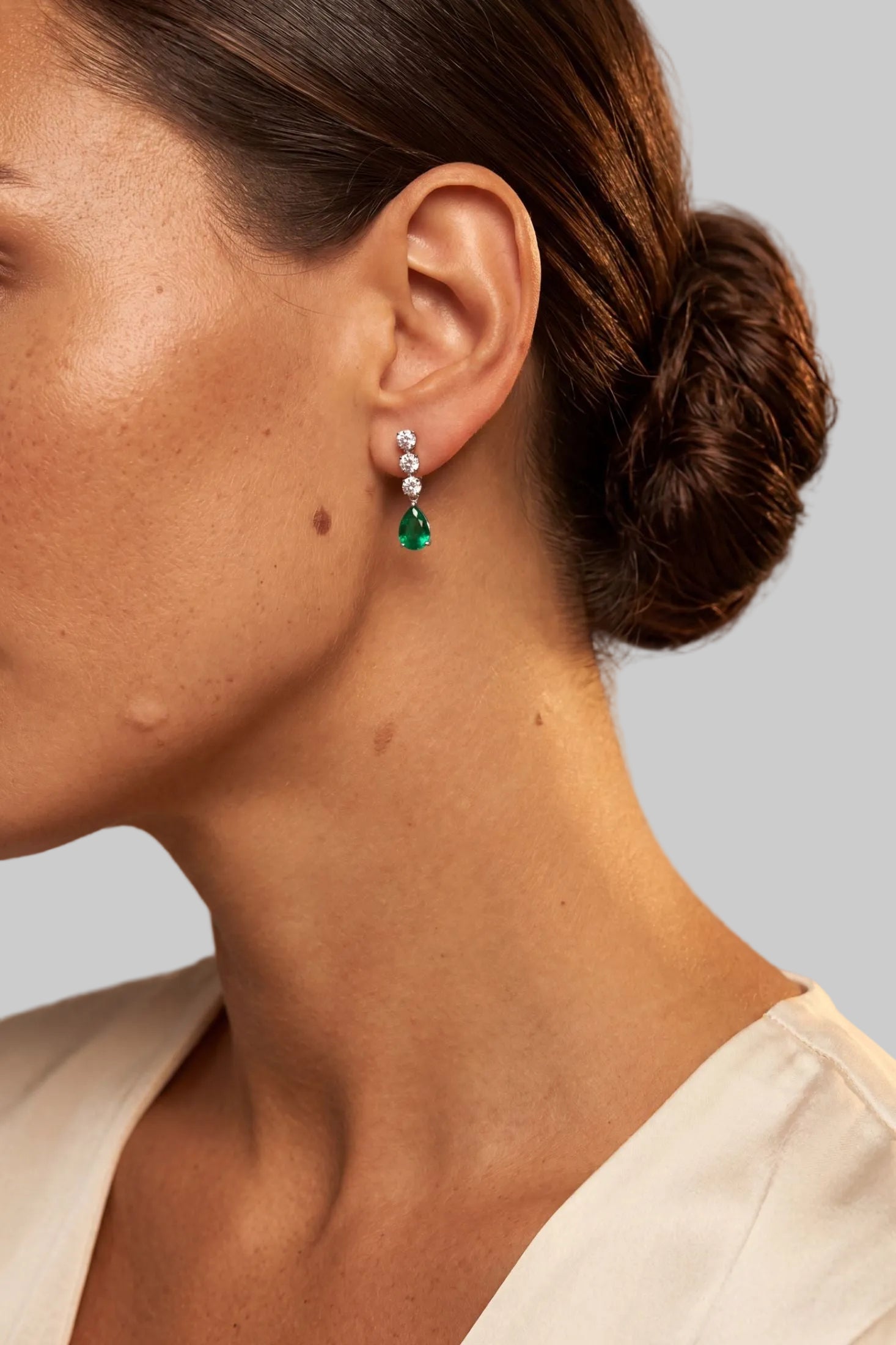 A close-up side profile of a woman wearing a single drop earring with a small silver-toned top and a green teardrop-shaped pendant, against a soft neutral background.