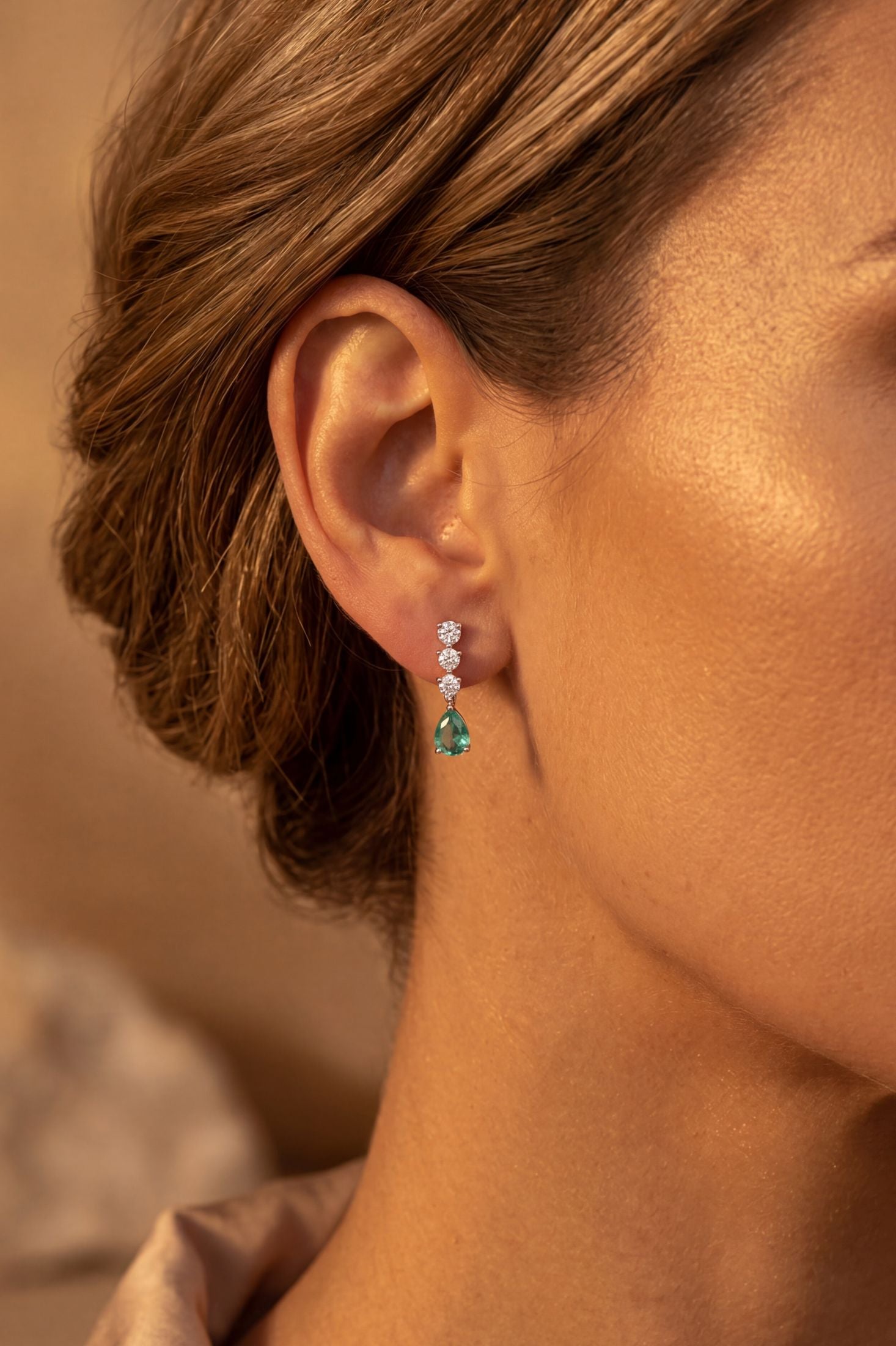A close-up side profile of a woman wearing a delicate drop earring with small clear stones and a green teardrop-shaped gemstone, set against a warm, softly blurred background.