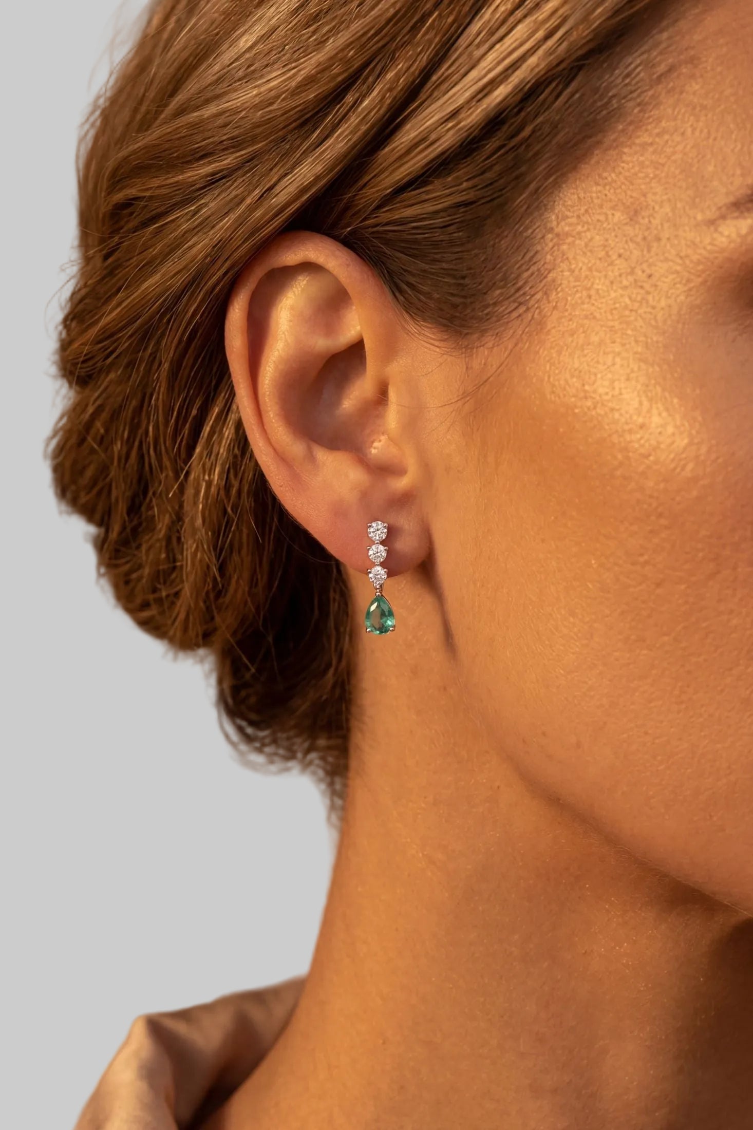 A close-up side profile of a woman wearing a delicate drop earring with small clear stones and a green teardrop-shaped gemstone, set against a warm, softly blurred background.