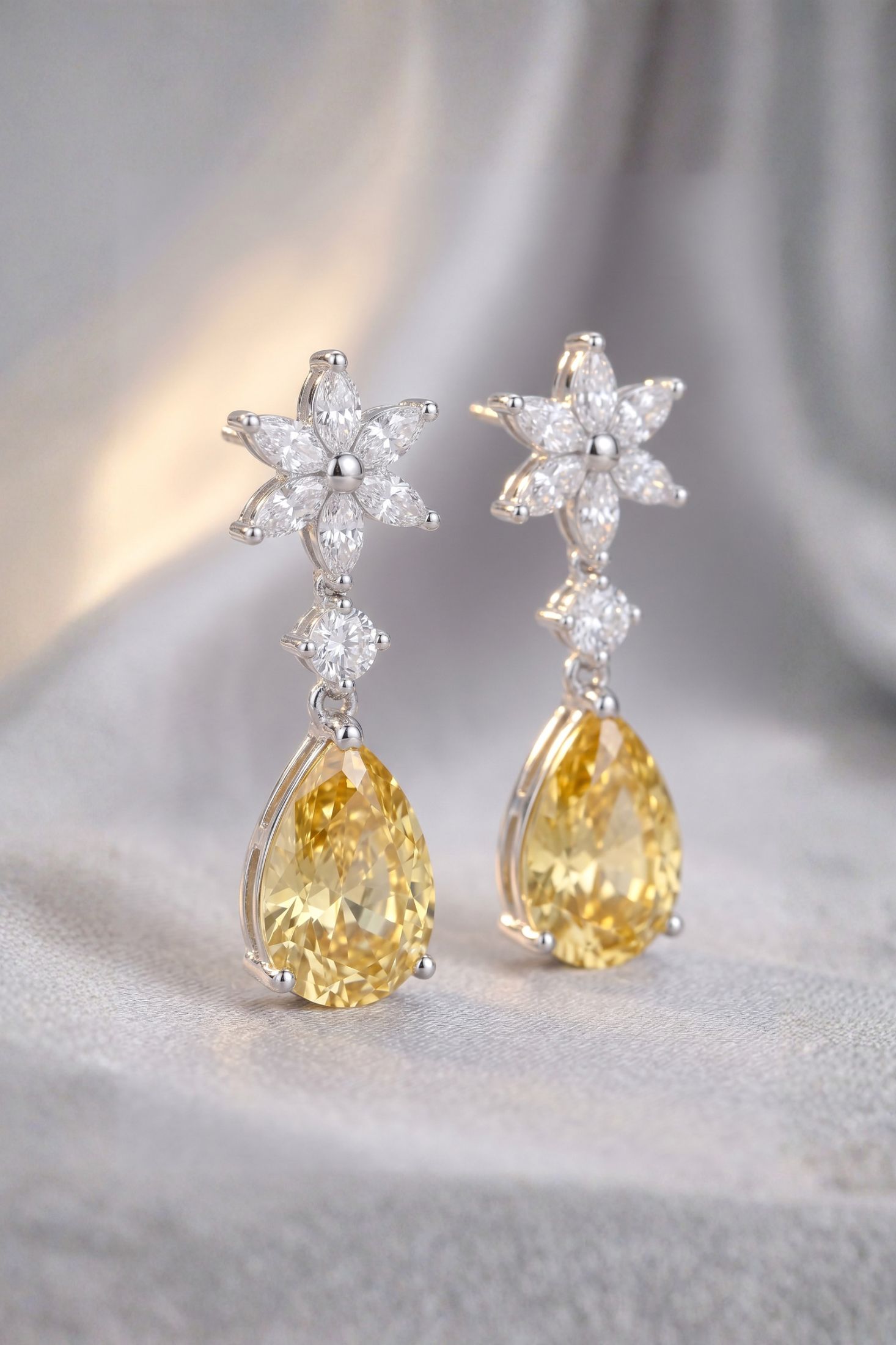 A pair of silver-tone dangle earrings featuring flower-shaped clusters of clear stones and teardrop-shaped yellow gemstones, displayed on a soft gray fabric background.