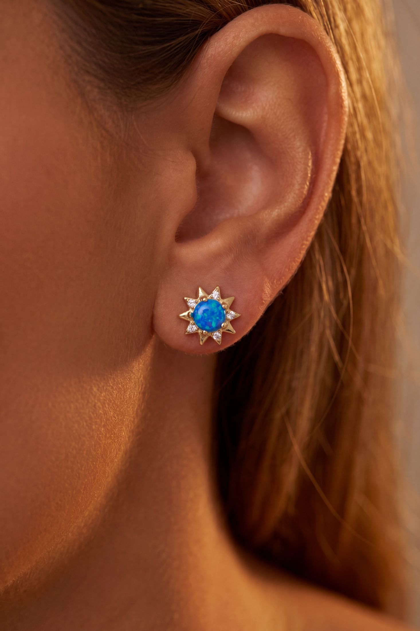 A close-up of a model's ear wearing a gold starburst stud earring with a vibrant blue oval center stone.