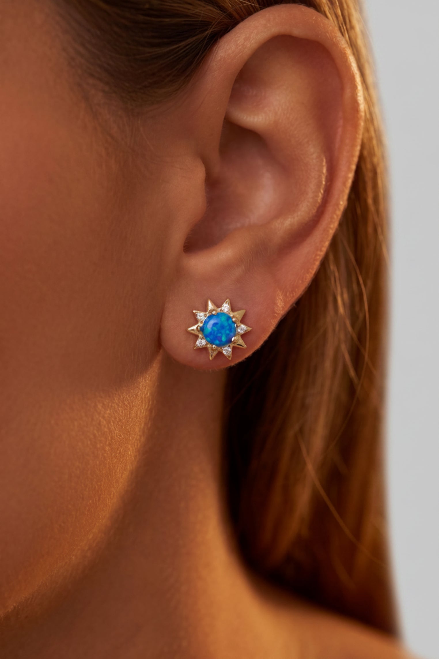 A close-up of a model's ear wearing a gold starburst stud earring with a vibrant blue oval center stone.