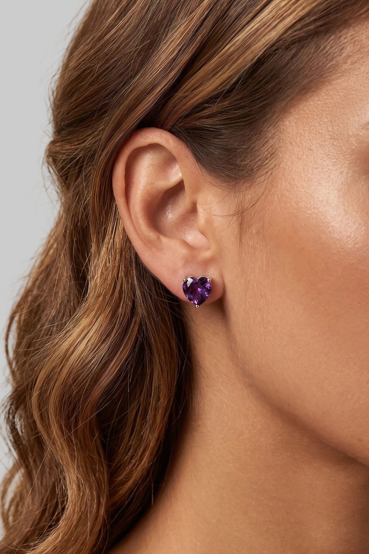 A close-up of a woman's ear wearing a small heart-shaped purple gemstone stud earring.