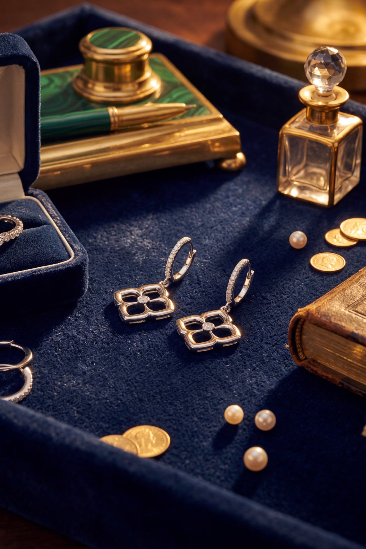 Silver four-leaf clover drop earrings displayed on a dark blue velvet jewelry tray, surrounded by vintage gold coins, a green and gold pen in a box, and a glass perfume bottle.