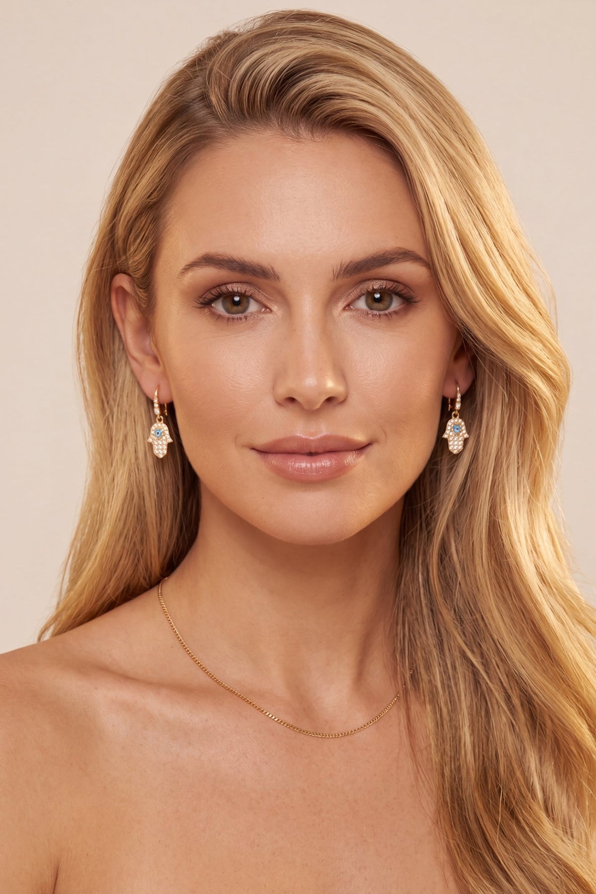 A close-up portrait of a blonde woman wearing dangling earrings with clear teardrop-shaped stones in a gold-toned setting, along with a delicate gold necklace, against a soft beige background.
