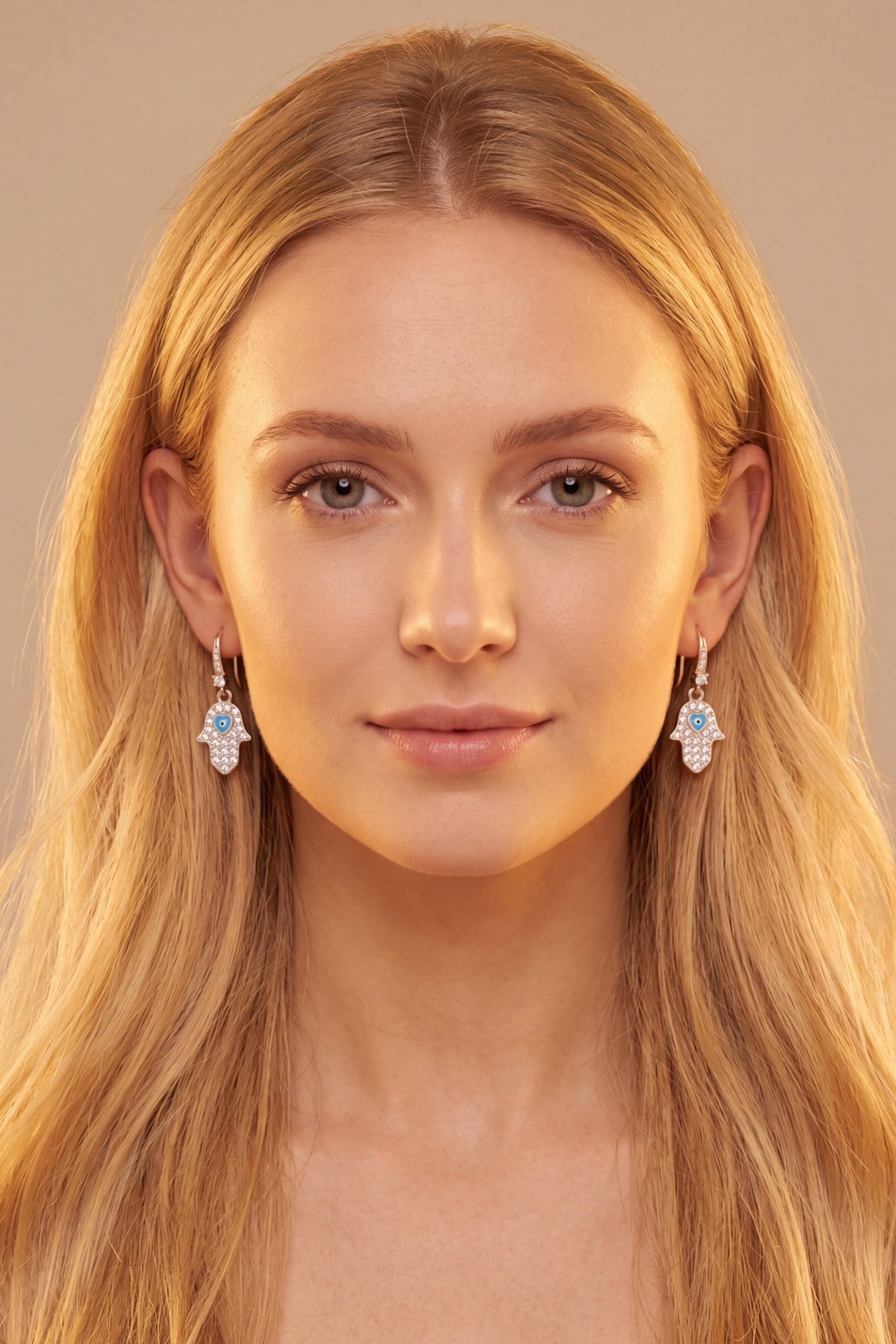 A close-up portrait of a woman with long blonde hair wearing delicate silver-toned dangle earrings with clear stones, set against a soft beige background.