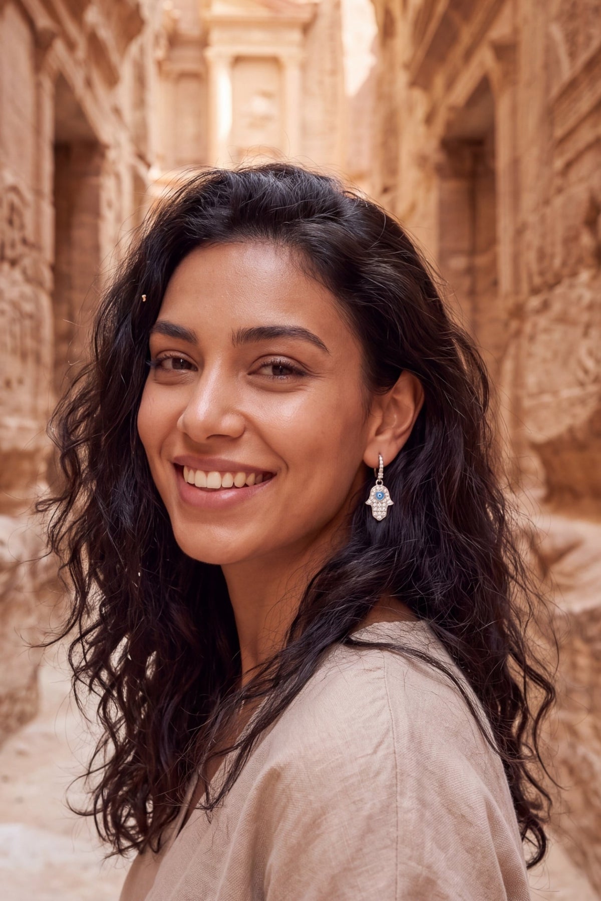 Smiling woman wearing a Hamsa hand earring in an ancient sandstone city setting.