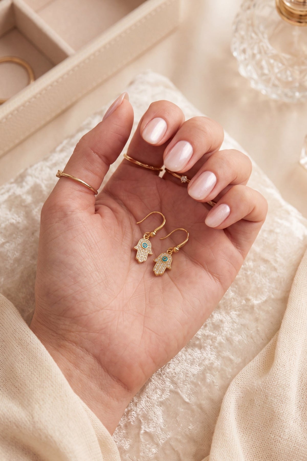 A close-up of a hand with pale pink manicured nails holding a pair of gold Hamsa hand earrings with small blue and green stone accents, styled on a beige textured fabric with jewelry boxes in the background.