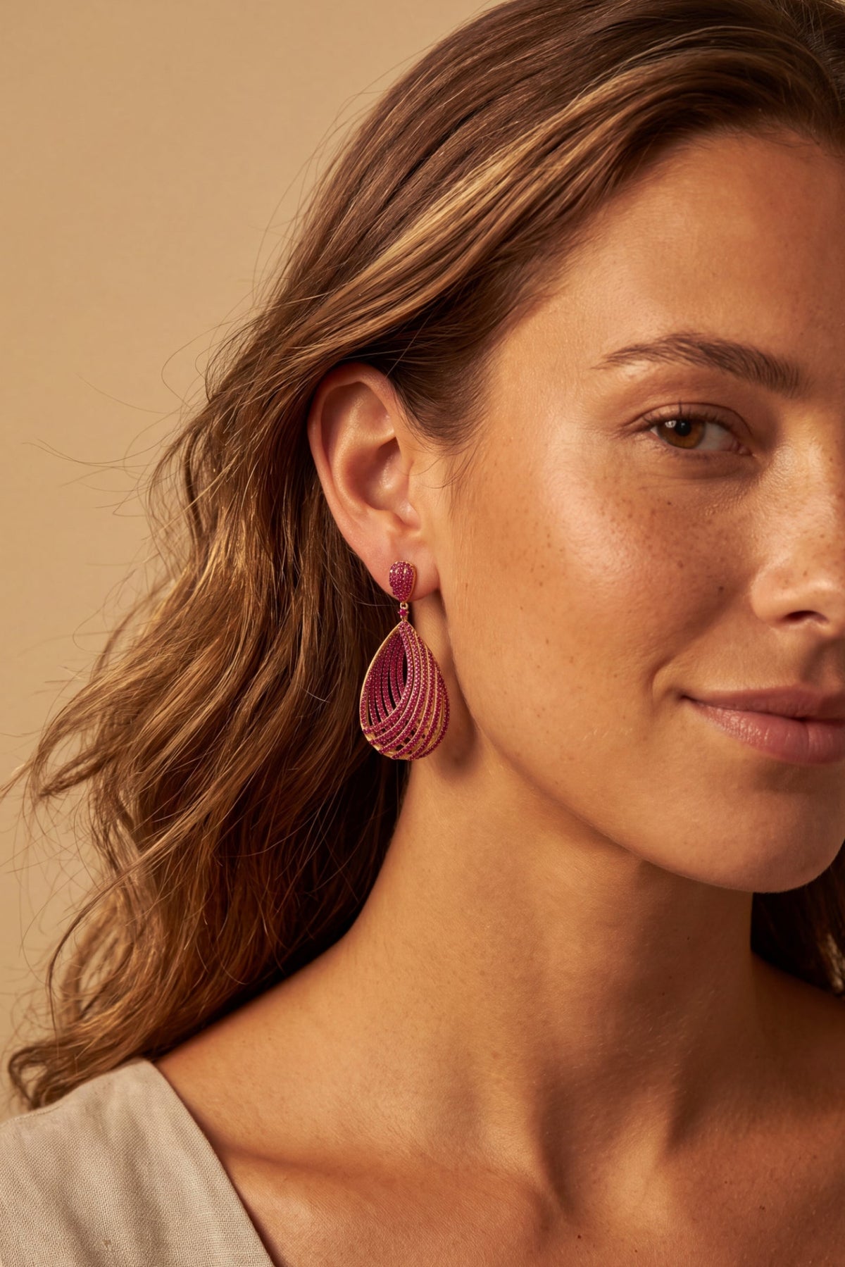 A close-up of a woman wearing a pink teardrop-shaped dangle earring with a textured, layered design. The background is a solid warm beige color.