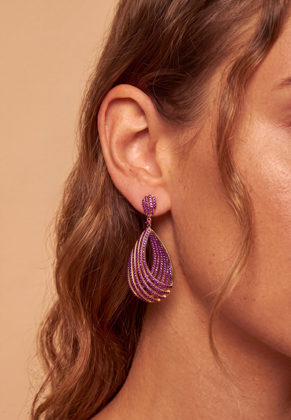 A close-up of a woman's ear wearing a teardrop-shaped dangle earring made of multiple rows of purple rhinestones with gold-toned metal edges and a gold-toned post.