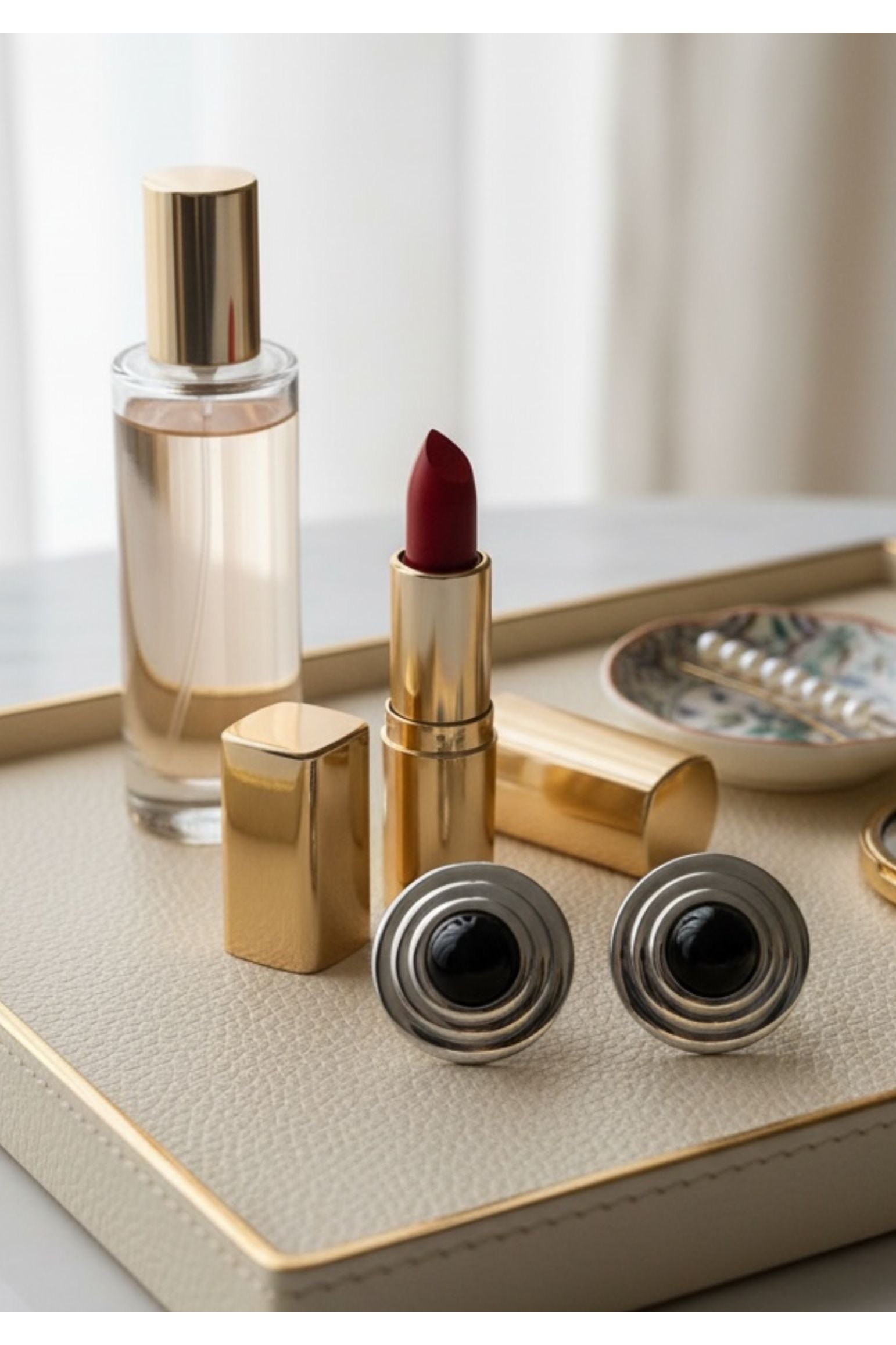 Gold lipstick, perfume bottle, and silver earrings with a circular black centre on a light surface with a neutral background