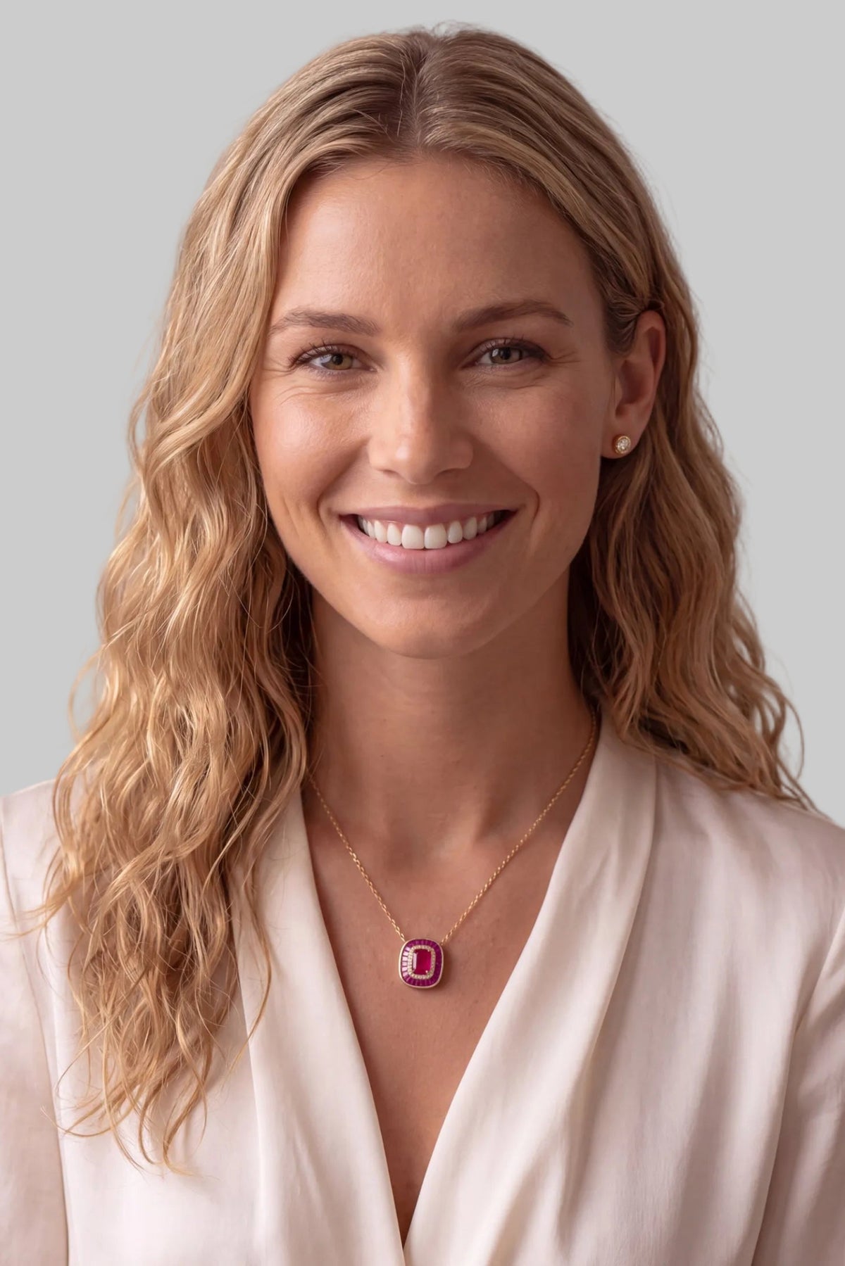 A smiling blonde woman wearing a delicate gold necklace with a rectangular pink gemstone pendant, styled with a cream-colored blouse against a neutral beige background.