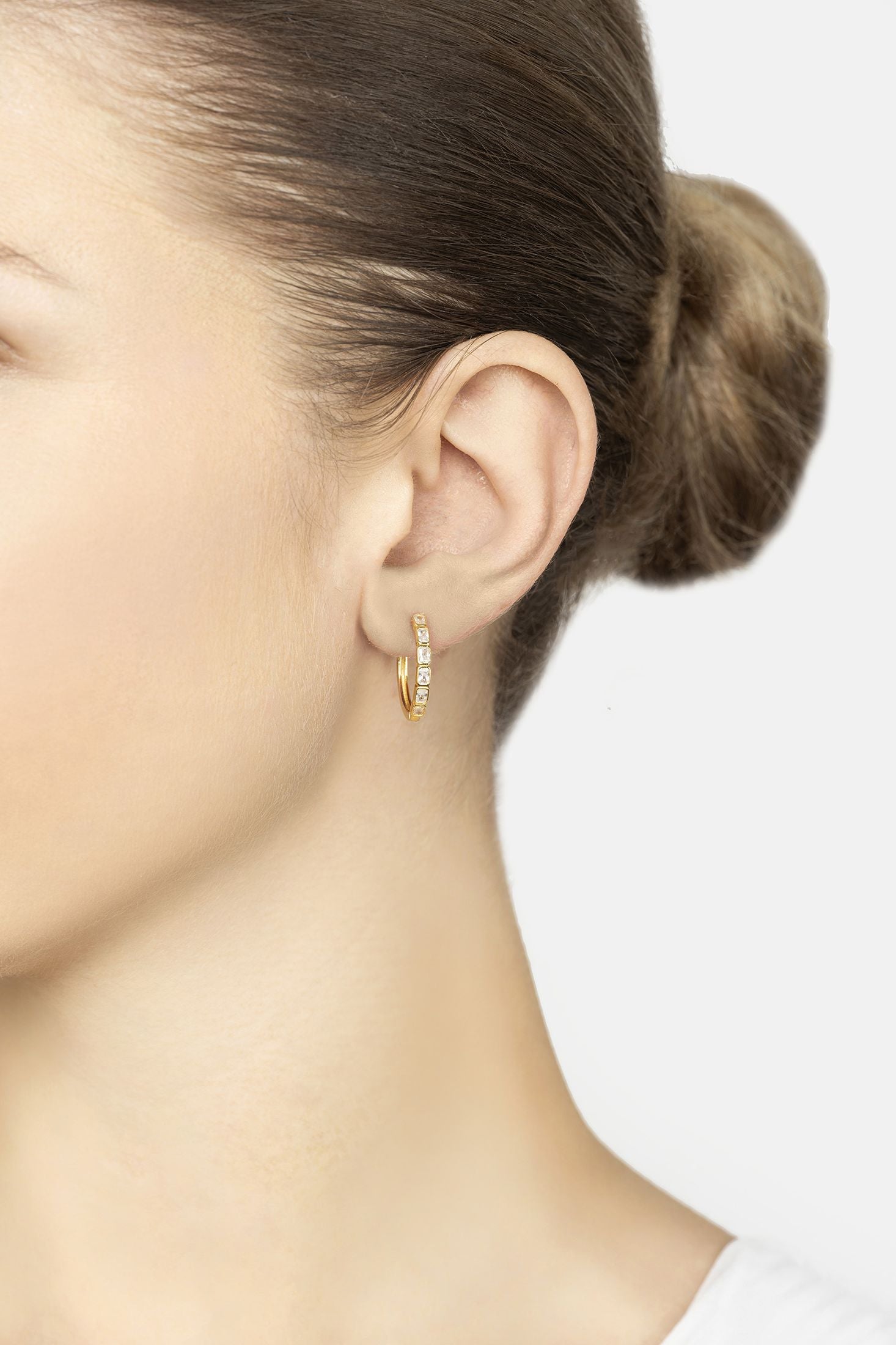 Small gold hoops with clear gemstones worn by a woman on a white background