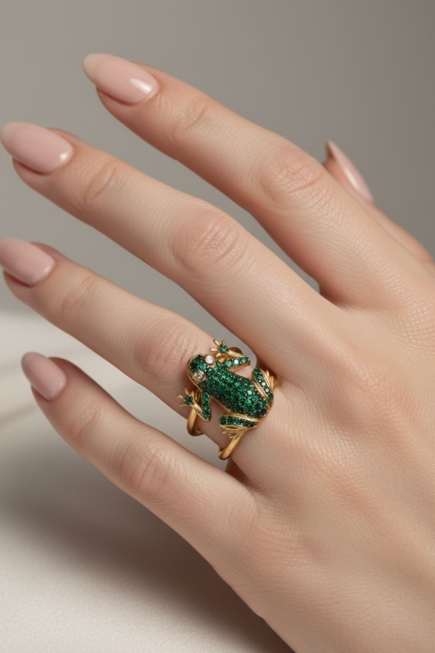 Hand wearing a gold frog ring with green gemstones on a neutral background
