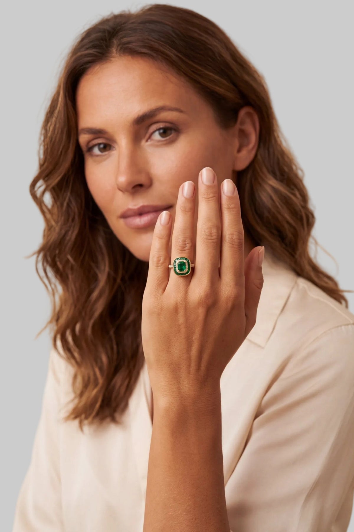 A woman wearing a silver ring with a green square-cut gemstone, holding her hand up to show the ring against a warm beige background.