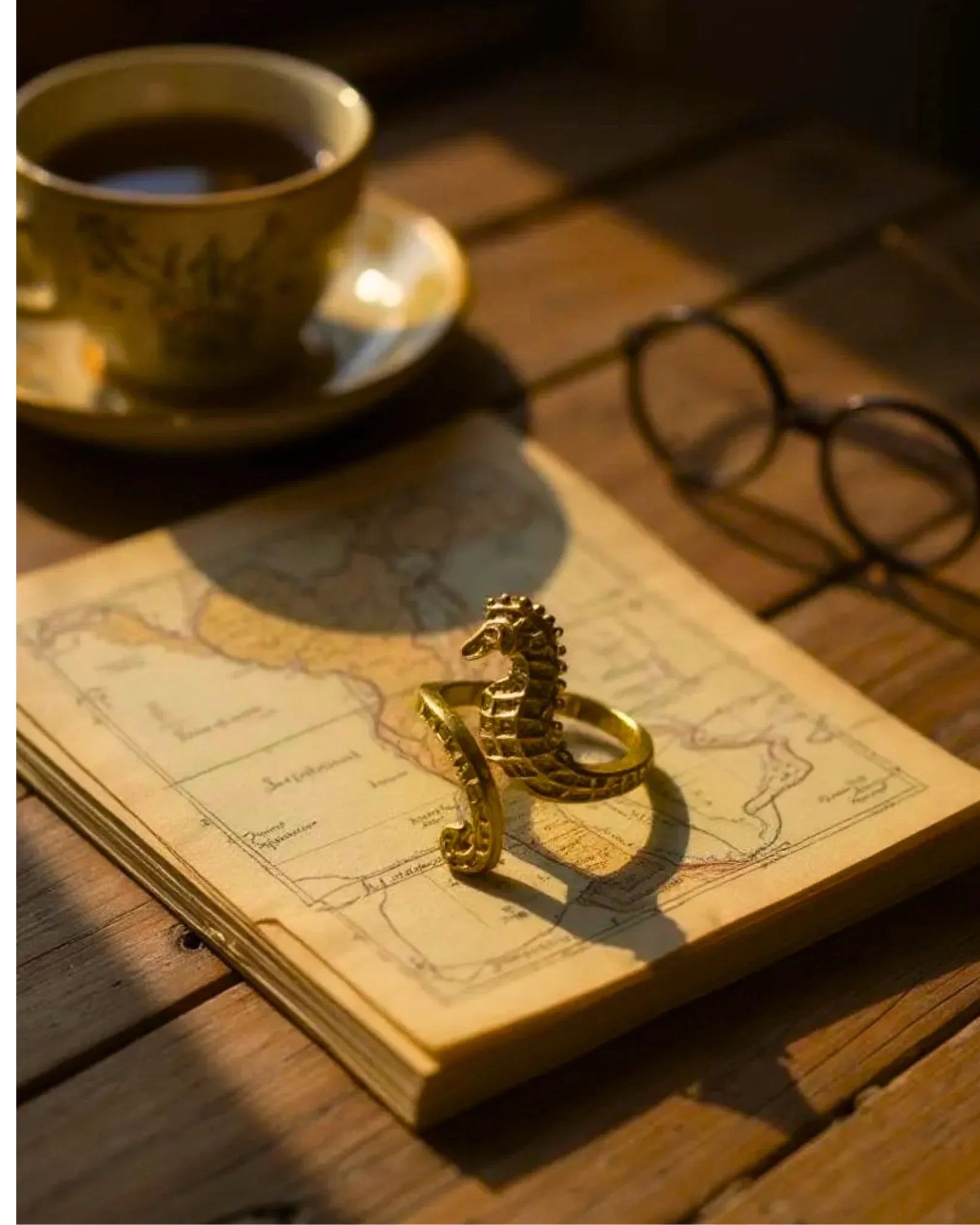 Gold seahorse ring on a pile of papers with a map on the top, a cup of tea and glasses on a wooden desk.