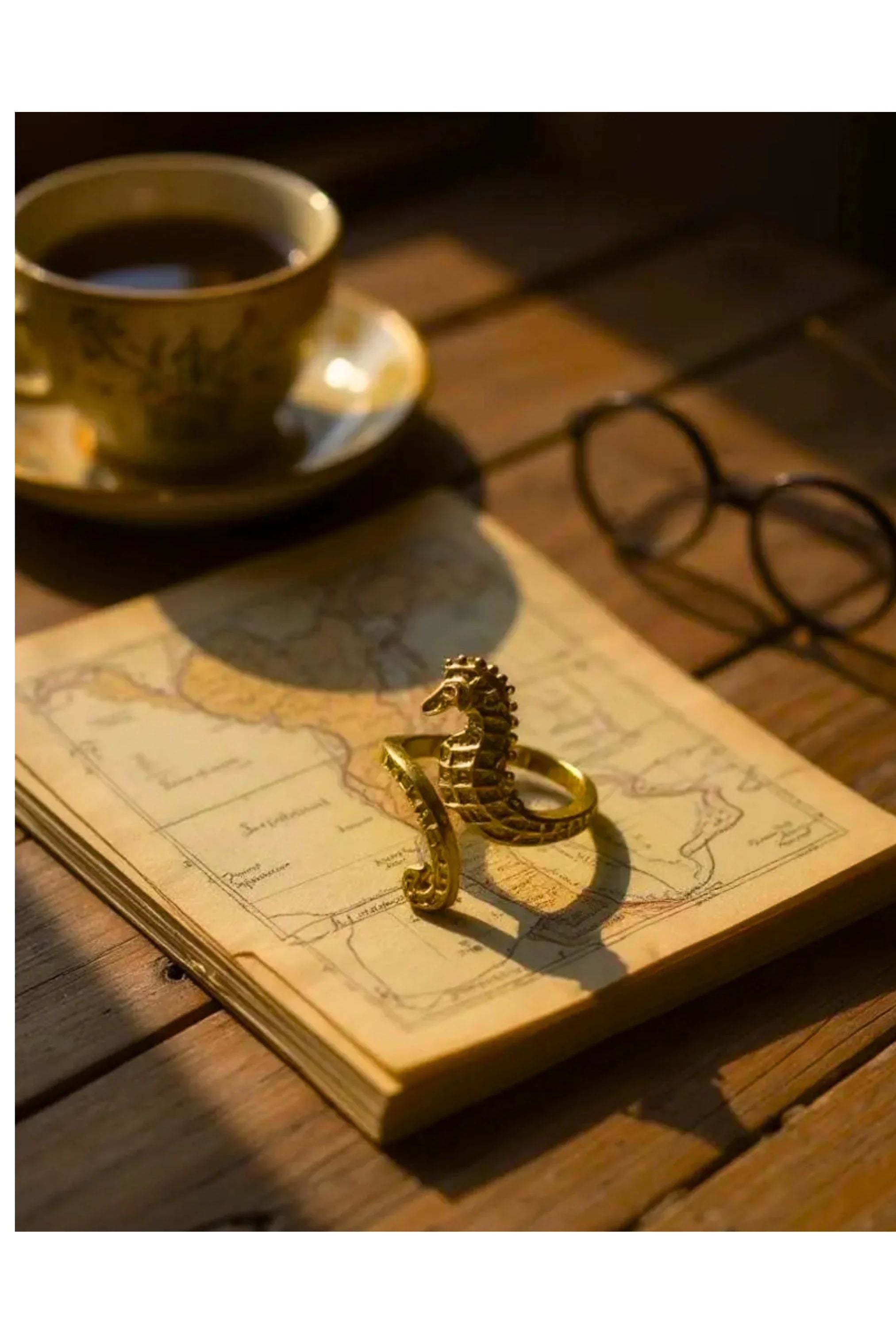 Gold seahorse ring on a pile of papers with a map on the top, a cup of tea and glasses on a wooden desk.