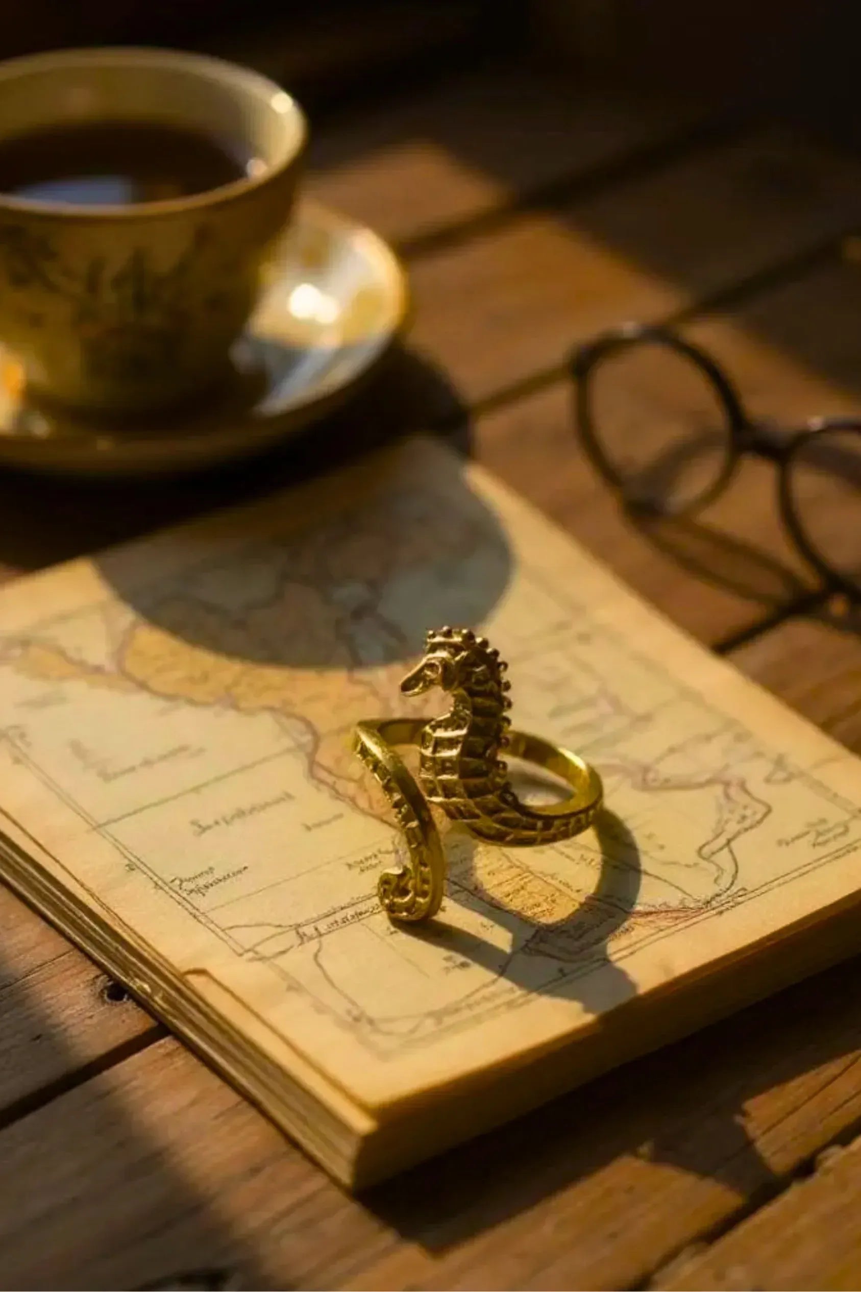 Gold seahorse ring on a pile of papers with a map on the top, a cup of tea and glasses on a wooden desk.