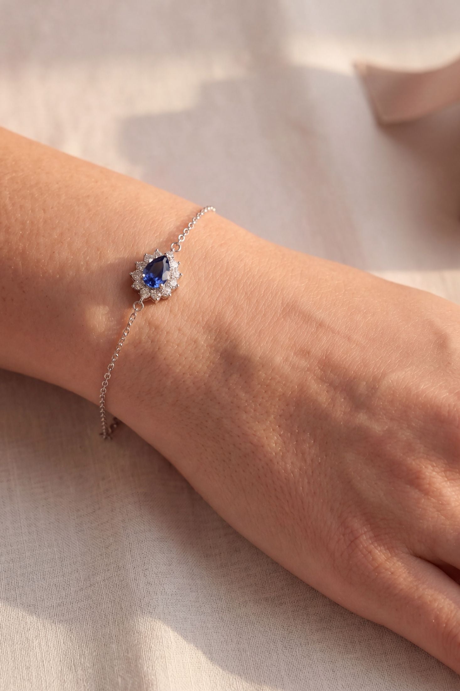 A delicate silver chain bracelet with a central teardrop-shaped blue gemstone surrounded by small clear stones, shown worn on a person's wrist against a soft beige fabric background.