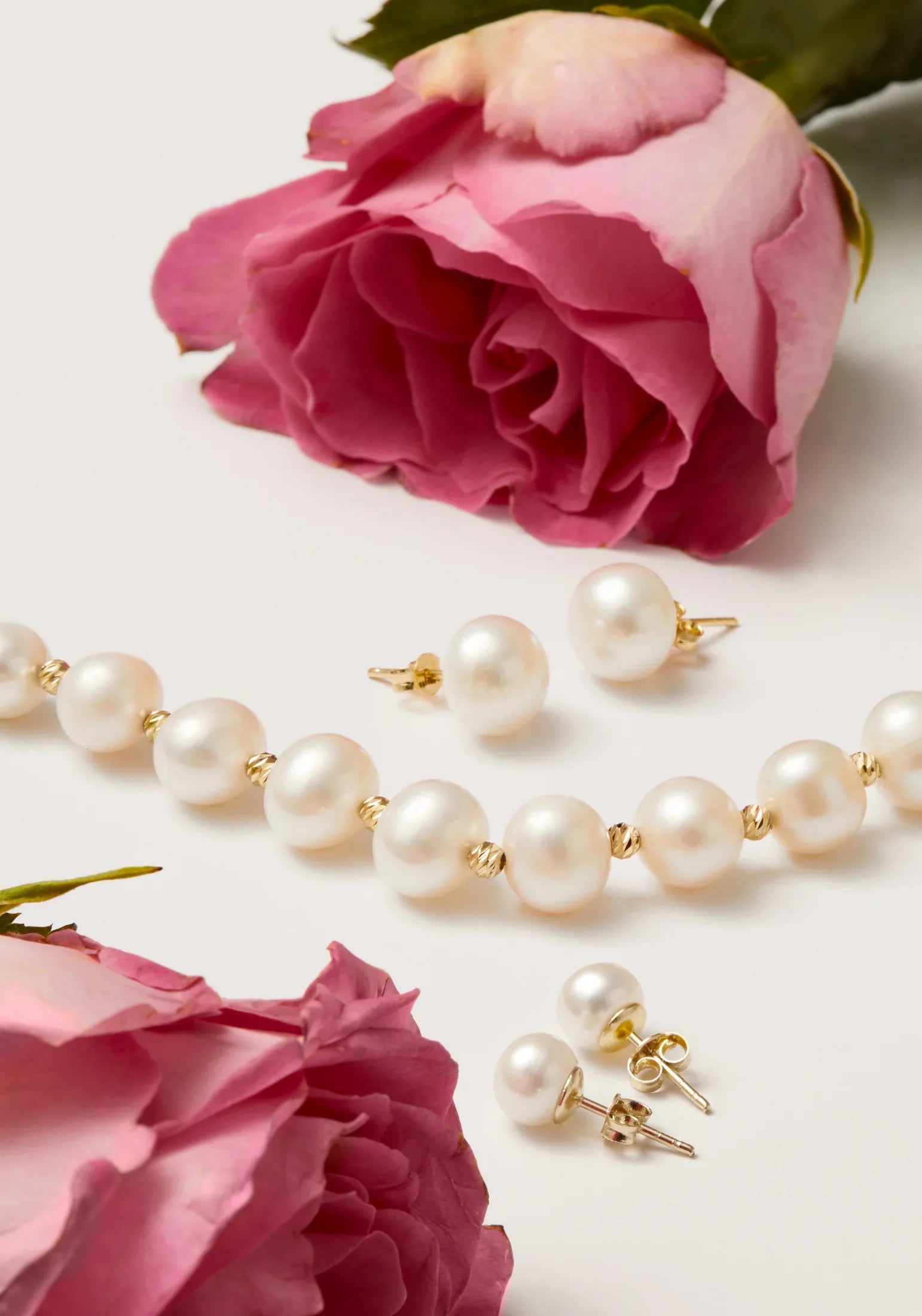 Pearl necklace and stud earrings alongside pink roses on a white surface.