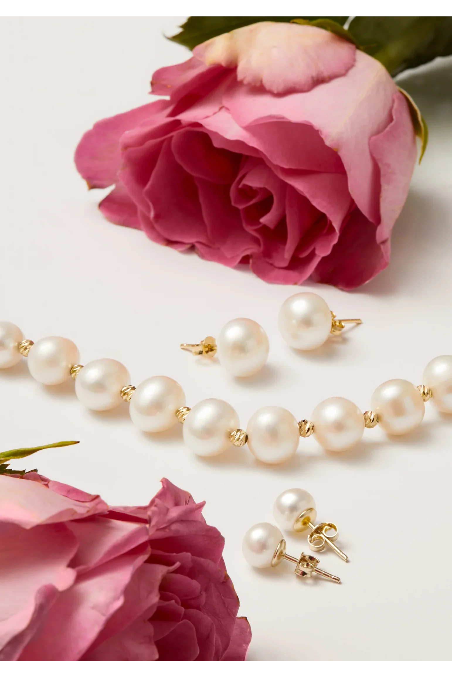 Pearl necklace and stud earrings alongside pink roses on a white surface.