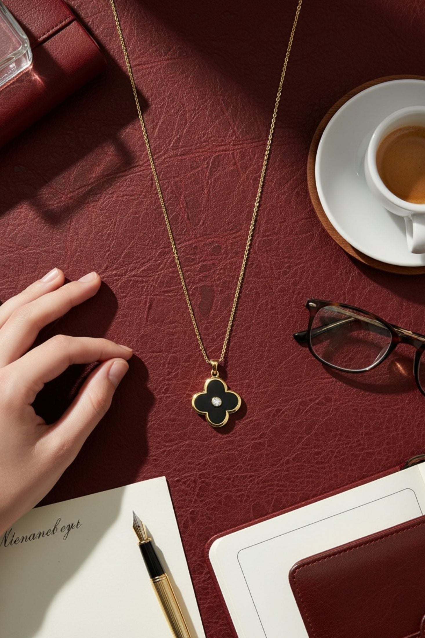 Gold necklace with a clover pendant on a red surface with a hand, glasses, and a cup.