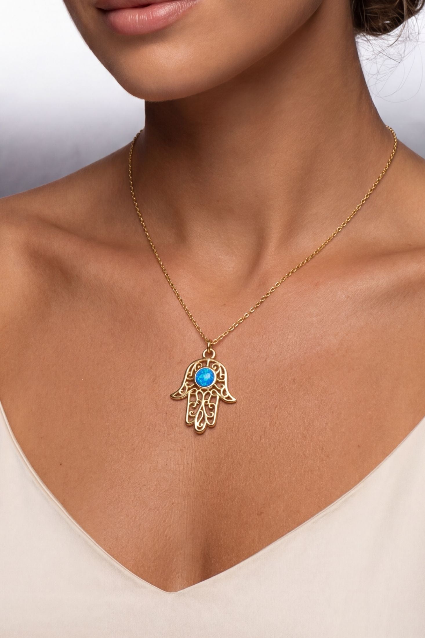 A gold Hamsa hand pendant necklace with a blue stone center, worn on a model's neck over a cream-colored top.