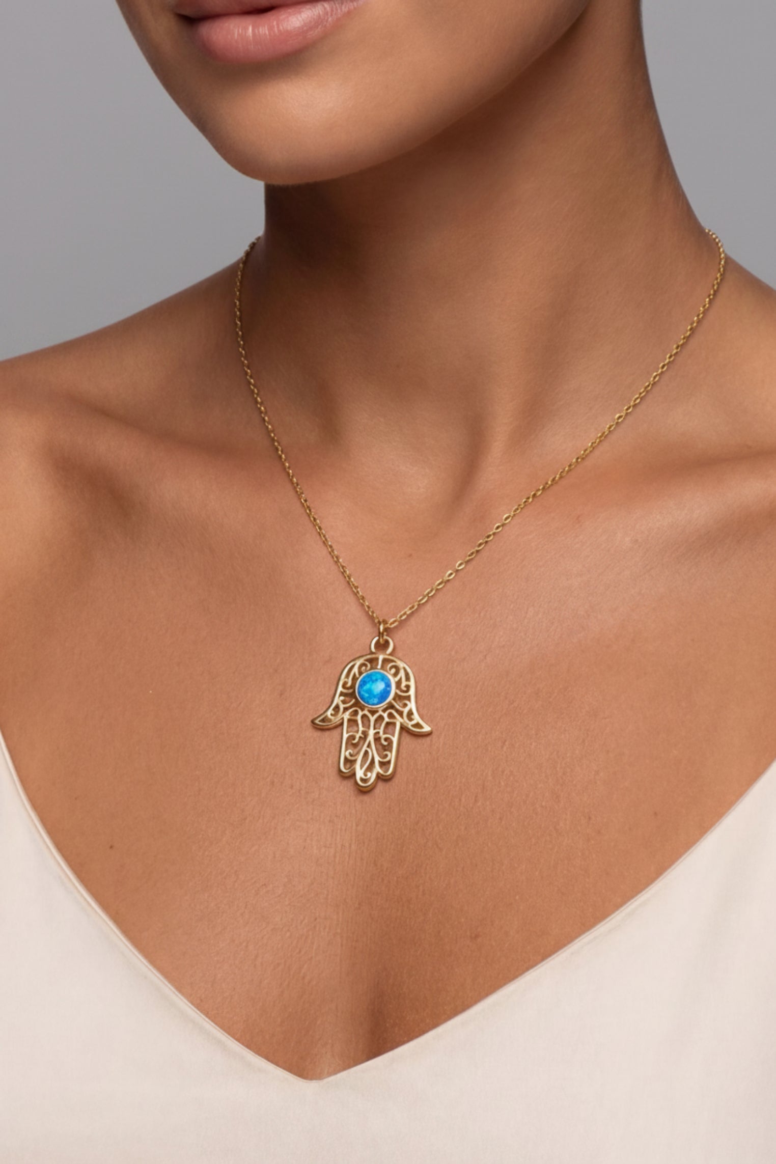 A gold Hamsa hand pendant necklace with a blue stone center, worn on a model's neck over a cream-colored top.