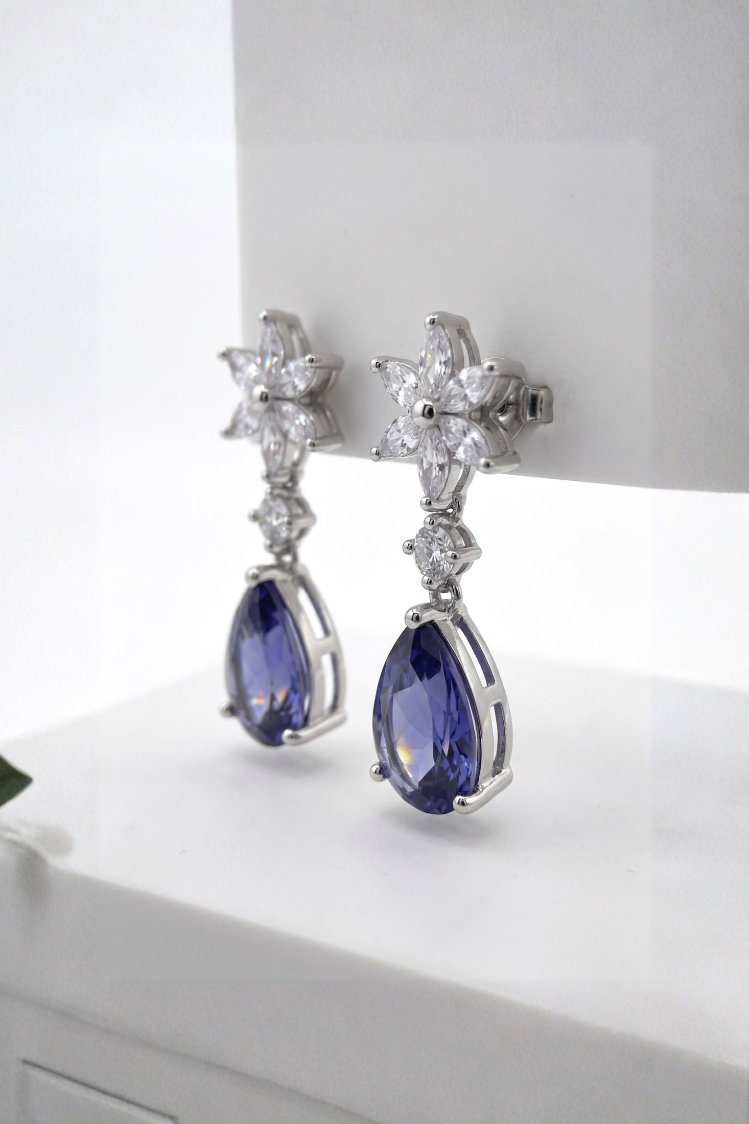 A pair of silver-toned drop earrings featuring starburst clusters of clear stones and pear-shaped deep blue gemstones, displayed on a white surface.