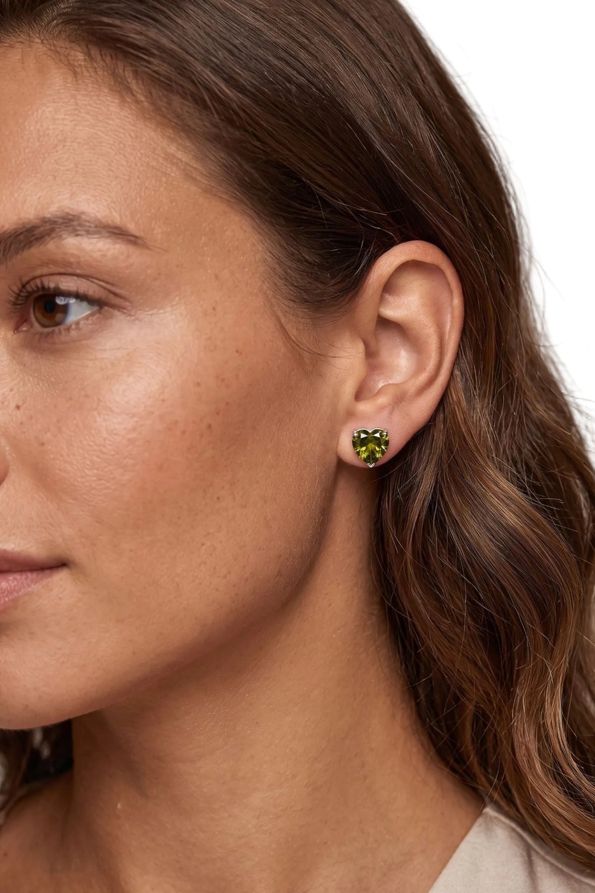 A close-up of a woman's ear wearing a small heart-shaped stud earring with a green heart in a gold setting.