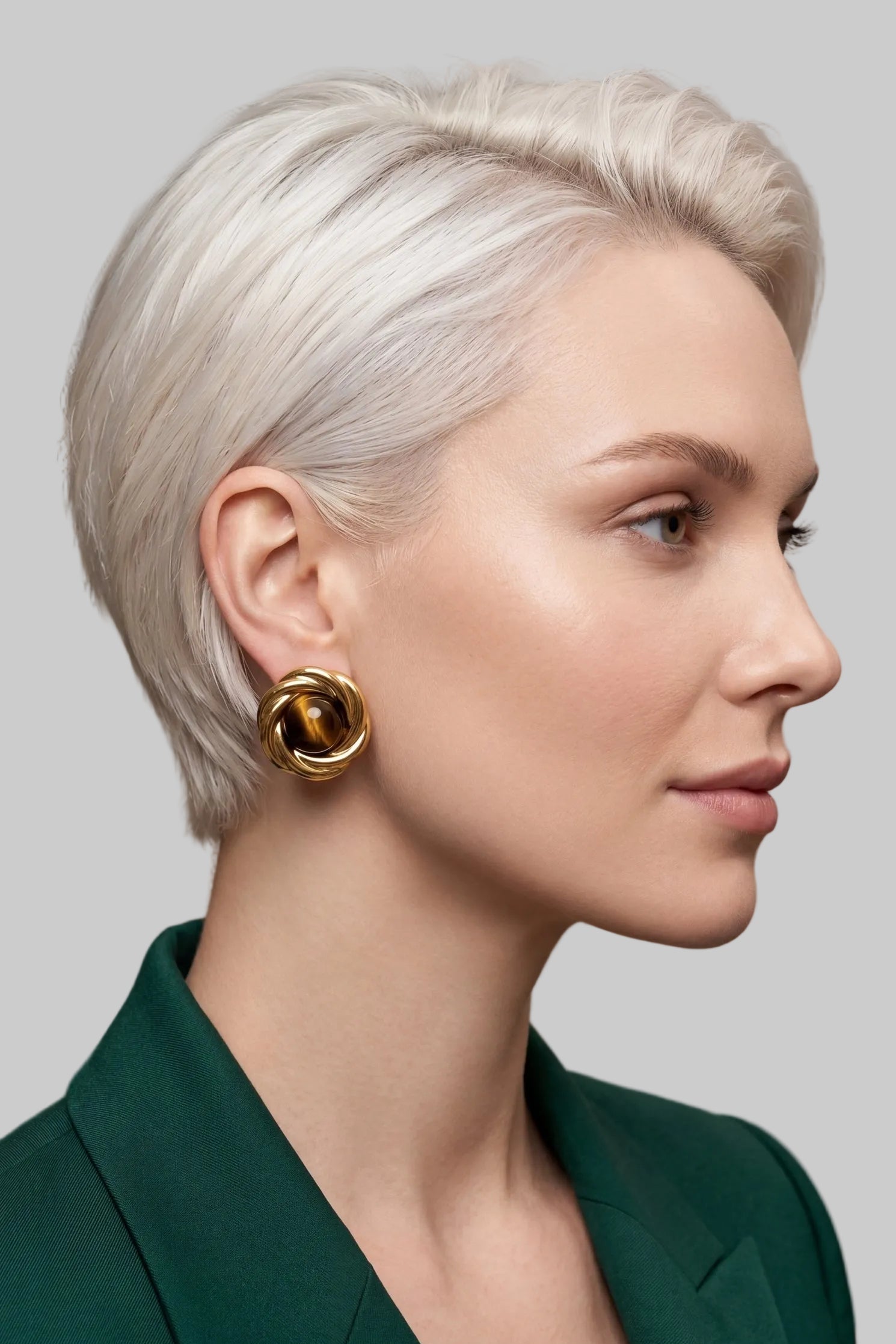 A close-up side profile of a woman with short platinum blonde hair wearing a gold geometric hoop earring and a dark green blazer.