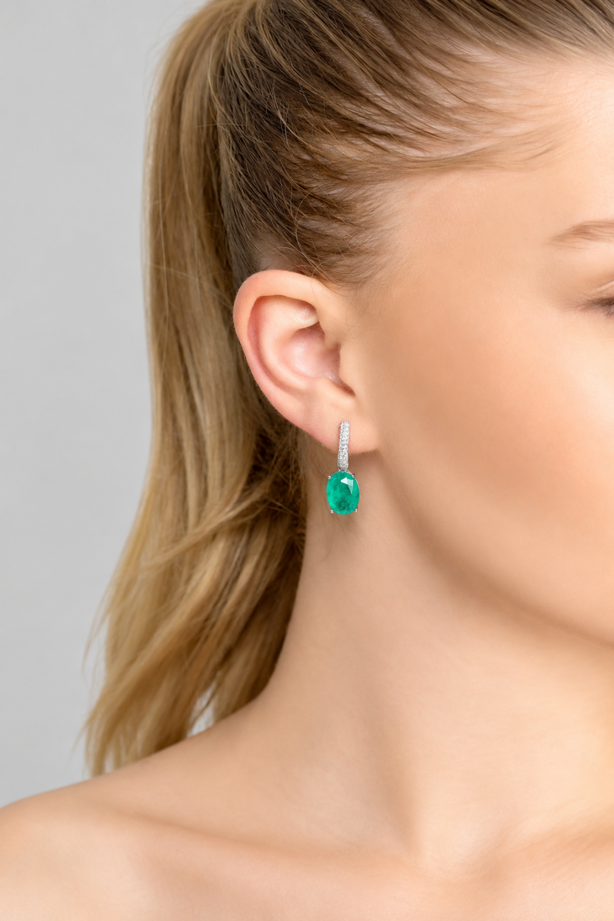 A close-up of a woman's ear and side profile wearing a single drop earring with a bright green oval gemstone and a silver-toned pavé setting.
