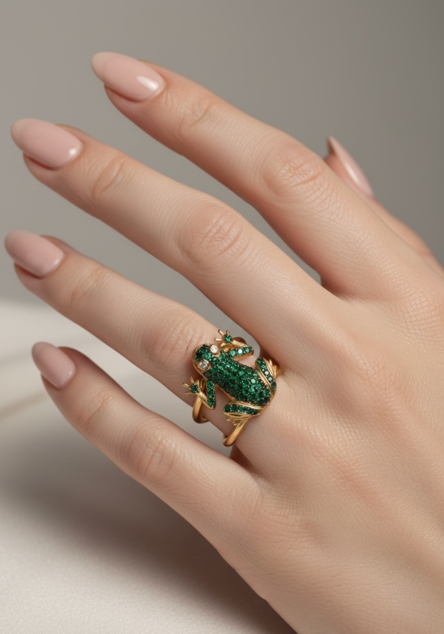 Hand wearing a gold frog ring with green gemstones on a neutral background